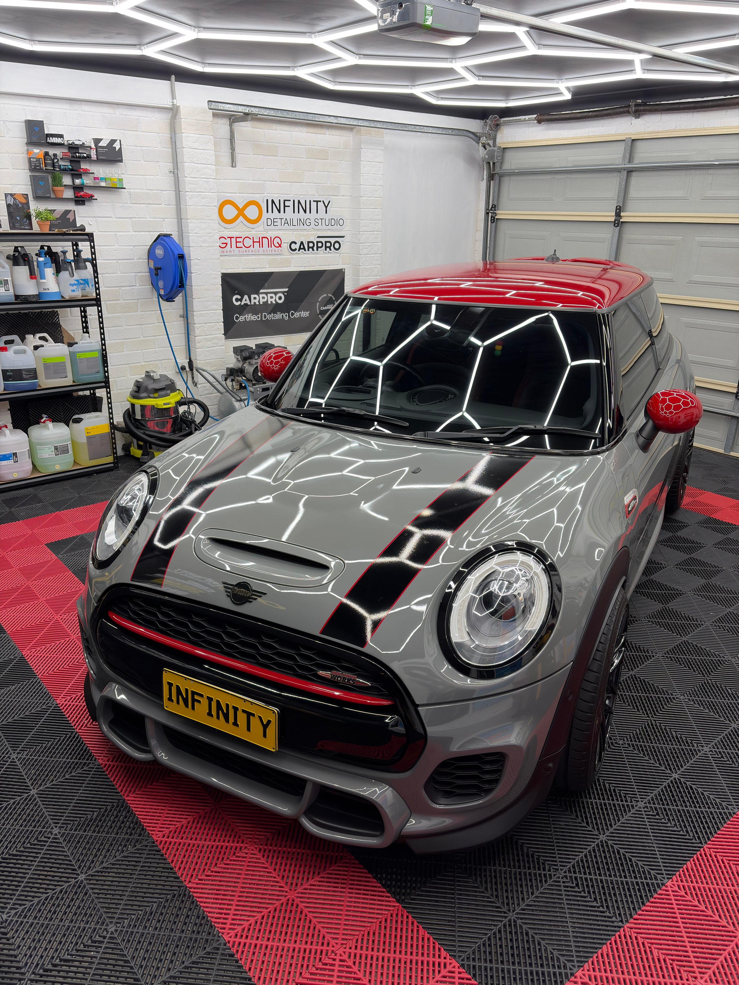 A Mini Cooper with a customized gray body, black racing stripes, received a paint correction. sign for Infinity Detailing Studio. The car is parked on a black and red checkered floor with futuristic lighting reflection