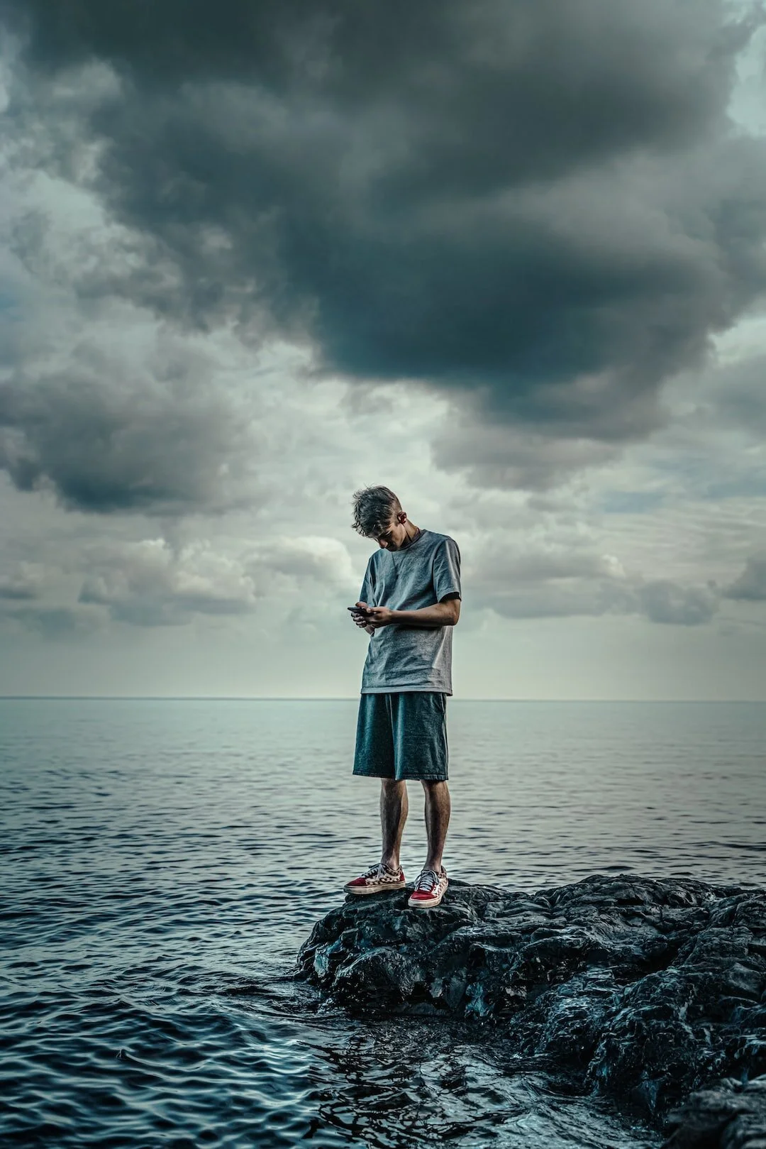 A young man standing on a rocky outcrop by the water, looking at his phone, with dark storm clouds overhead.