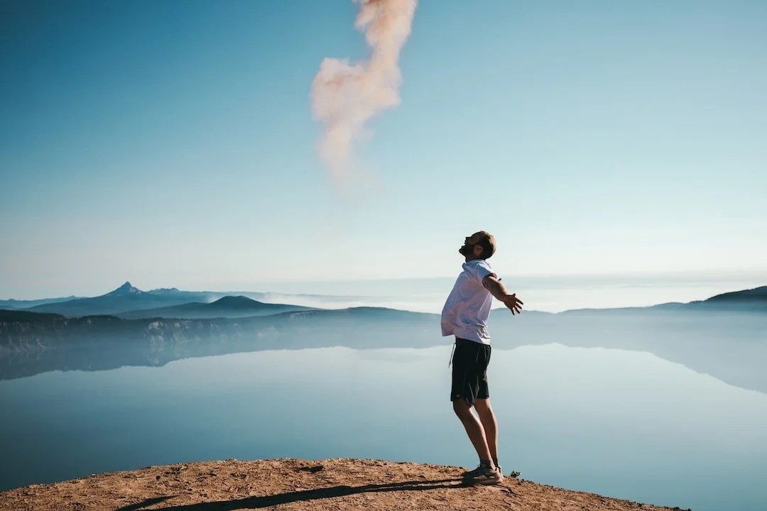 A person standing on a rocky cliff with arms open wide, facing a large body of water and distant mountains, with smoke rising in the sky.