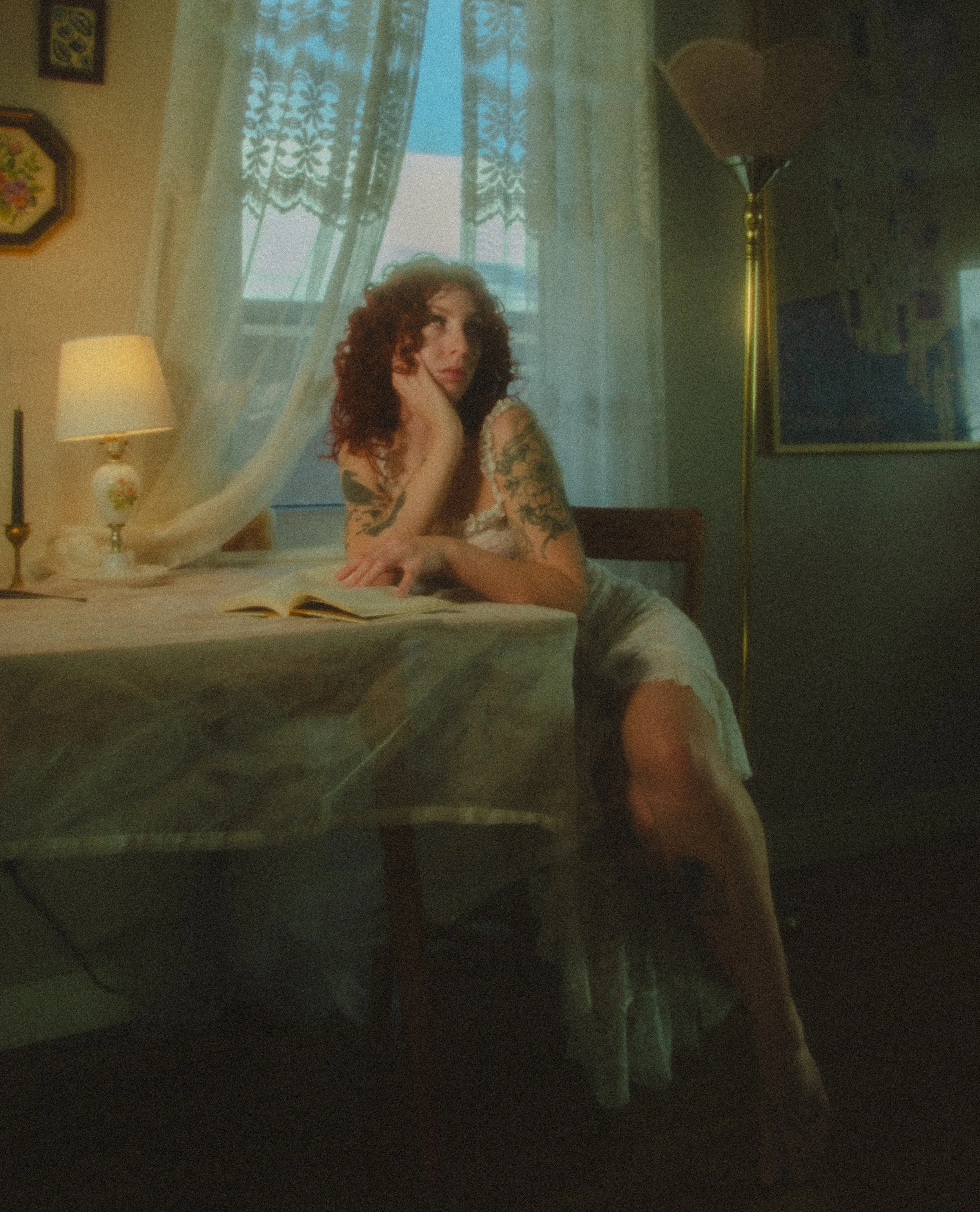 A woman with curly red hair and tattoos on her arms sitting at a table, resting her chin on her hand, in a dimly lit room with a lamp, framed artwork, and sheer curtains at the window.
