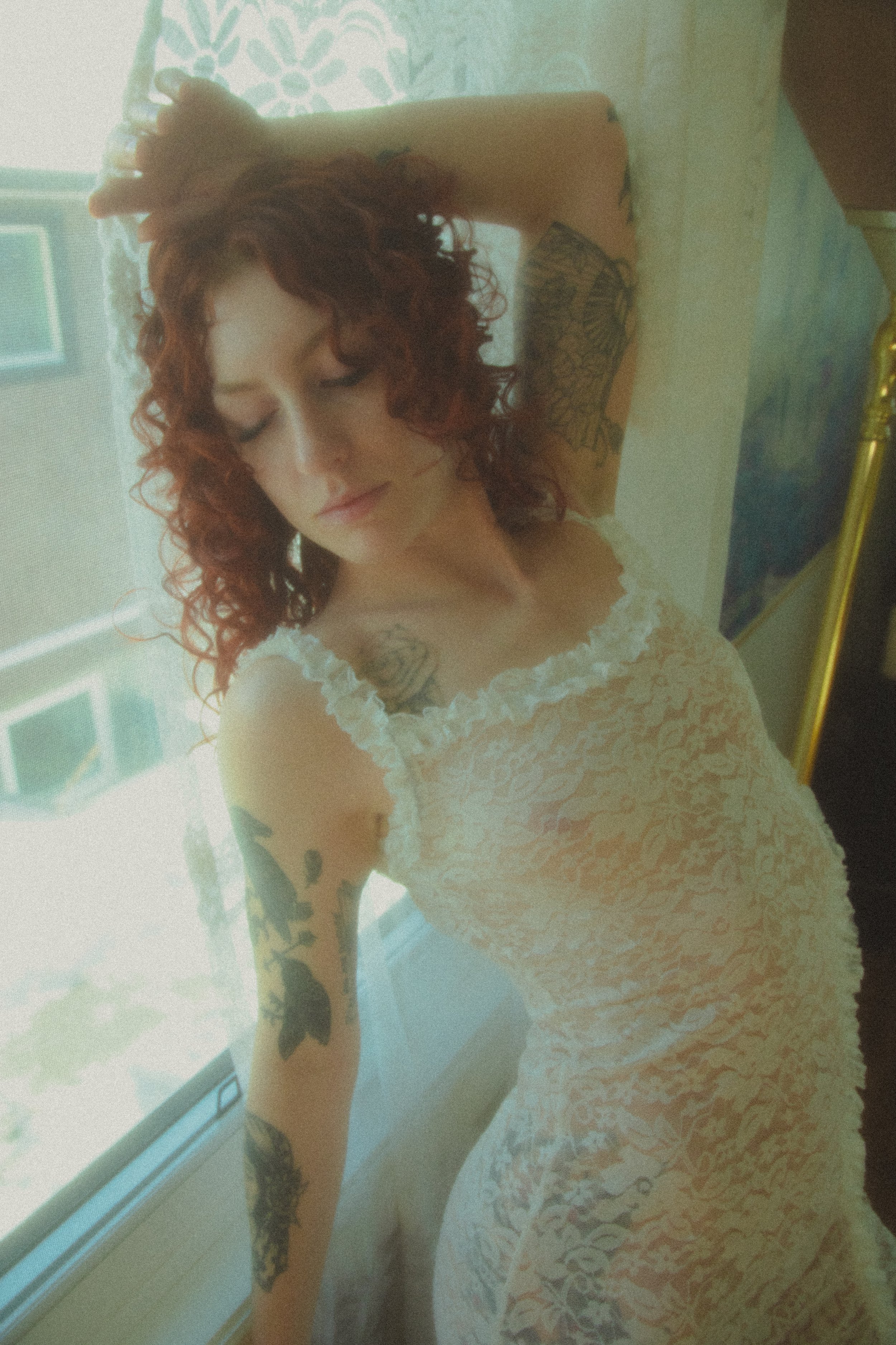 A woman with curly red hair and tattoos on her arm and chest, wearing a white lace dress, leaning on a windowsill with her eyes closed.