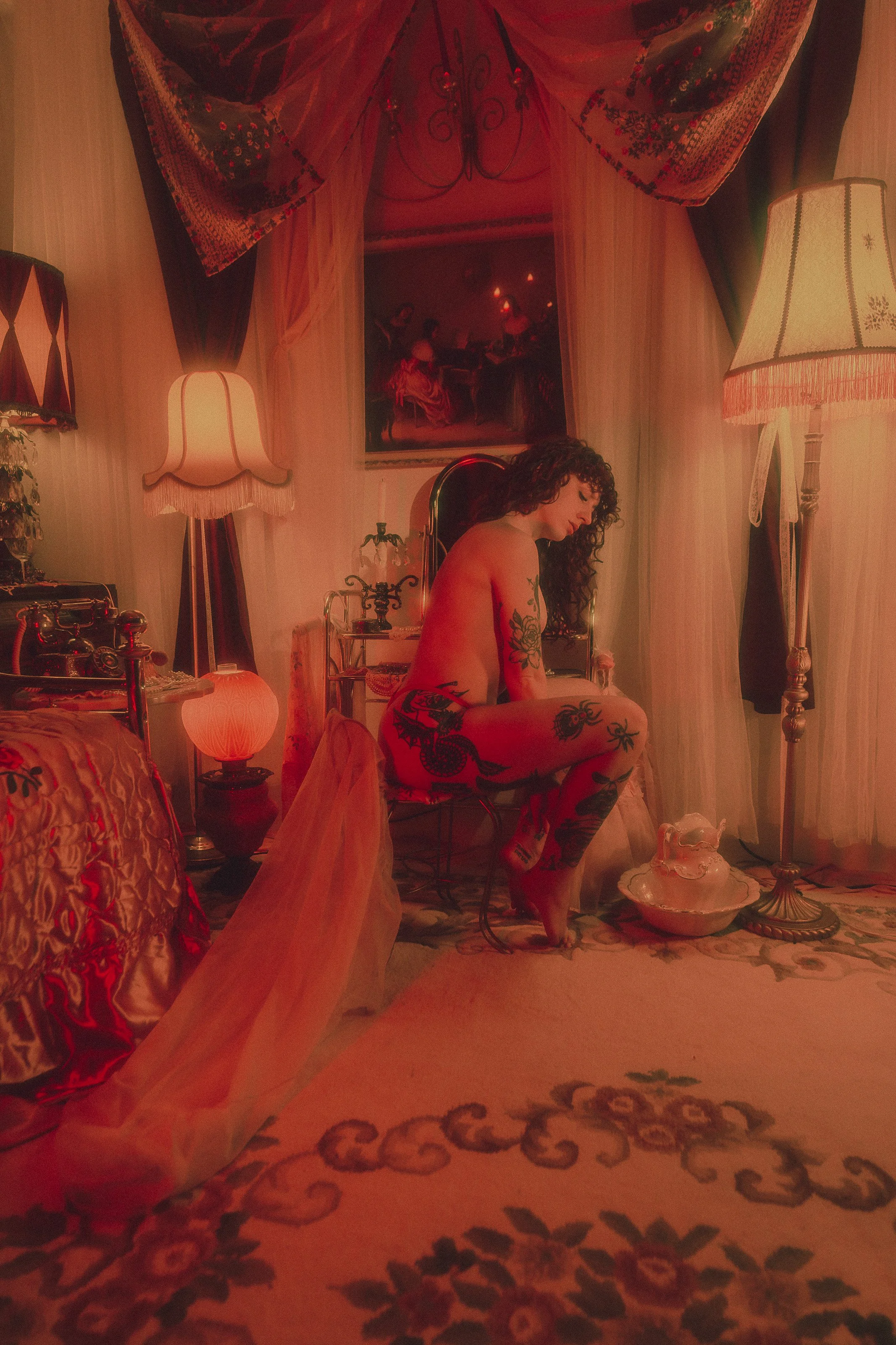 Image description: Jessie is in a valentine's themed room in front of a vanity, bathed in red light from the left and soft warm lamp light on the right. She sits facing left on a vintage metal framed chair, leaning slightly forward with her arms plac