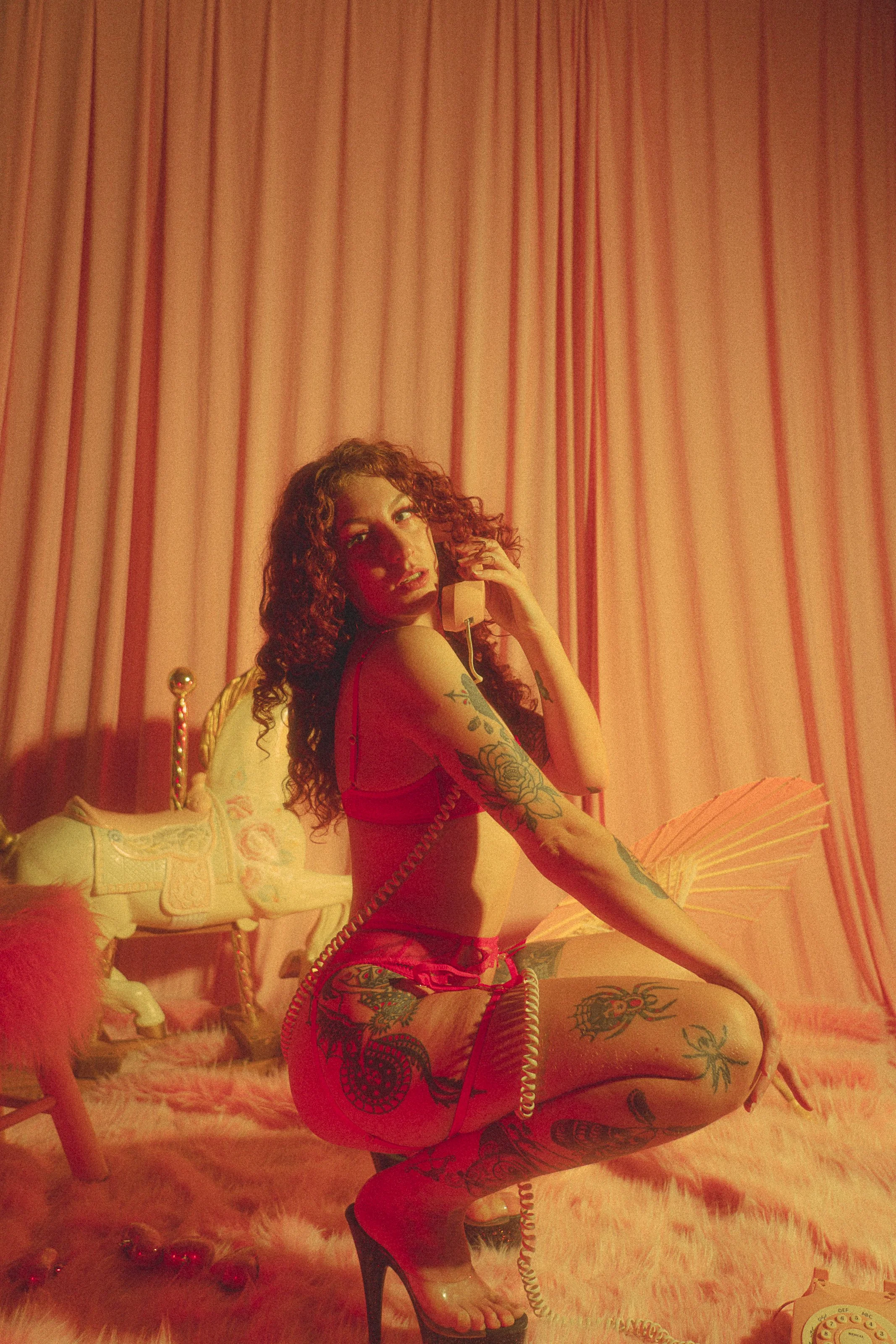 A woman with tattoos on her arms and legs, wearing red lingerie and high heels, sitting on the floor in a room with pink lighting, vintage style decor including a white rocking horse, pink curtains, and fluffy pink area rug, holding a yellow corded p