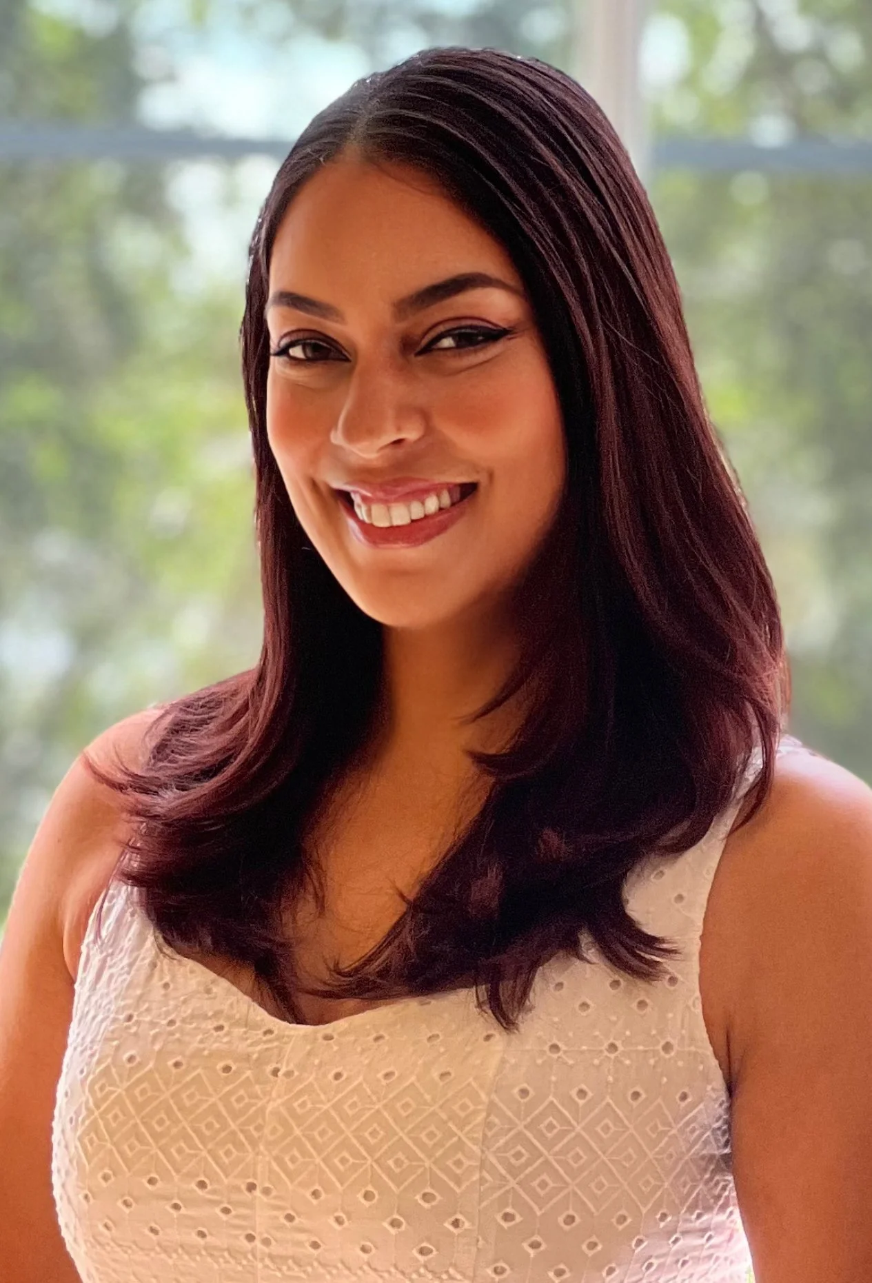 A woman with dark red, wavy hair, smiling and wearing a sleeveless white dress with eyelet details, standing in front of a blurred outdoor background.