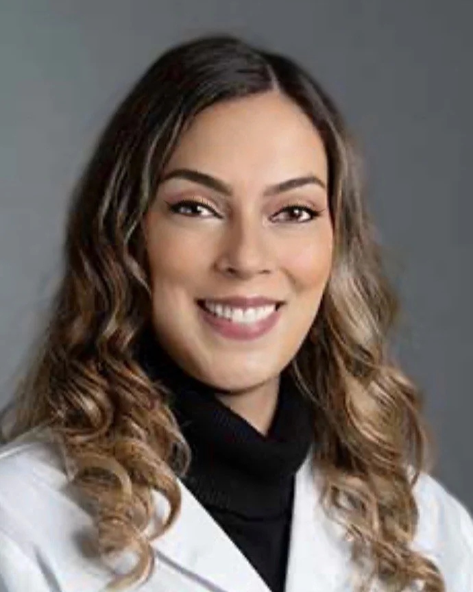 Headshot of a smiling woman with wavy light brown hair, wearing a white lab coat and a black top, against a gray background.