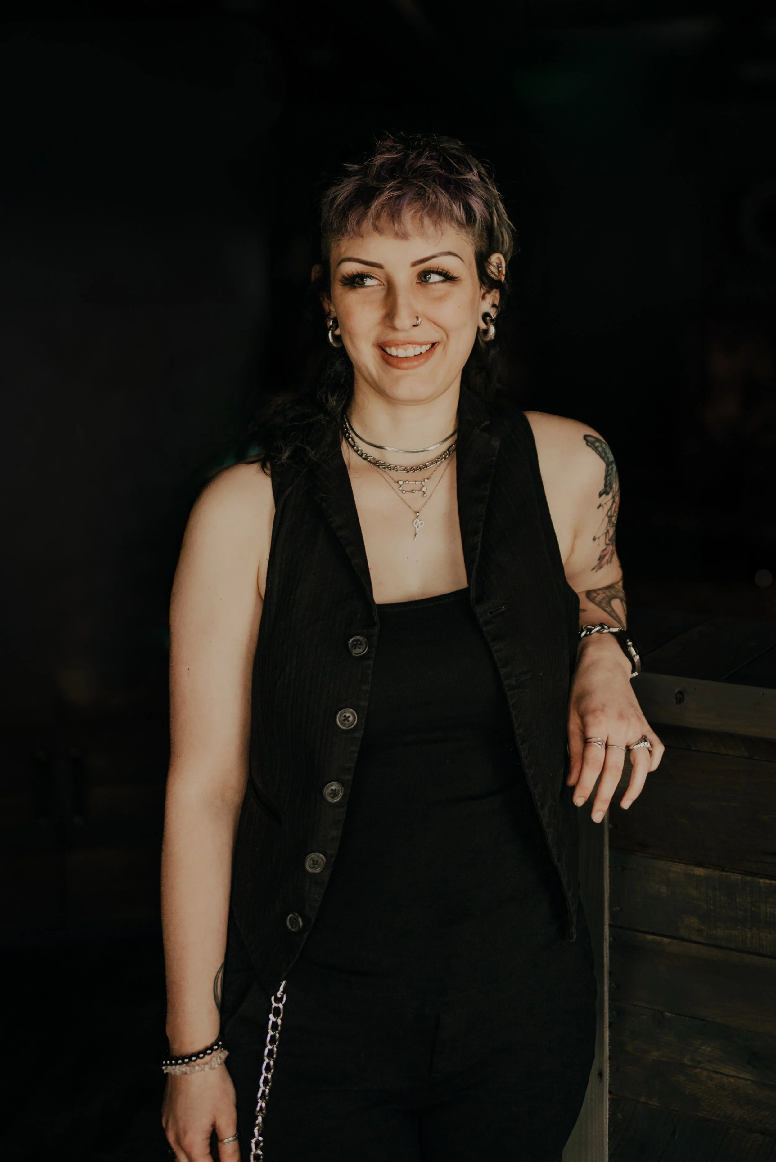 A hairstylist with a wavy hair mullet and tattoos on her upper arm, wearing layered necklaces and earrings. She is smiling and looking slightly to her left, dressed in a black sleeveless top, against a dark background. Alt vibes.