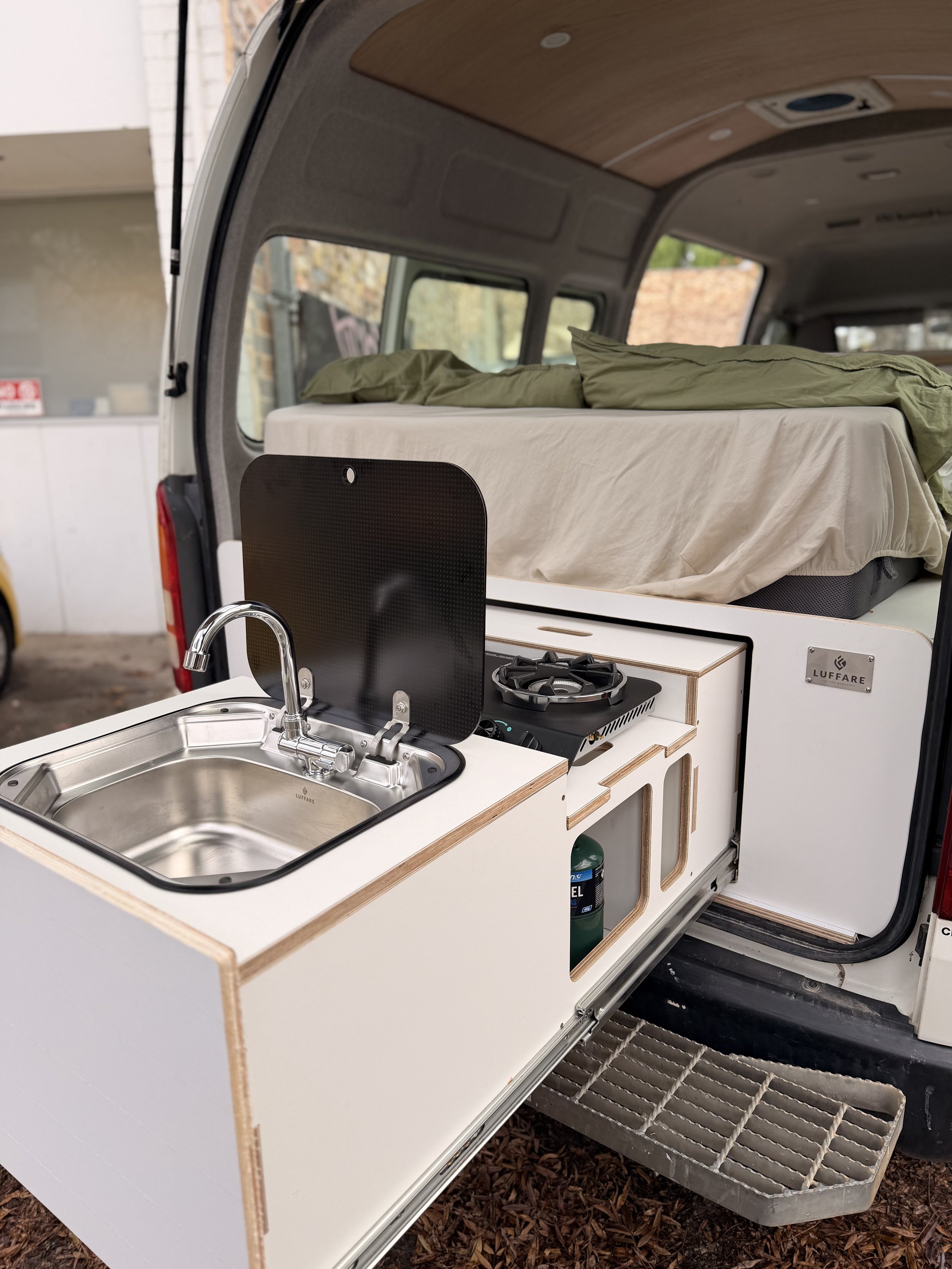 Interior of a compact camper van with a small kitchen area, featuring a sink and a two-burner stove, with a bed in the background.