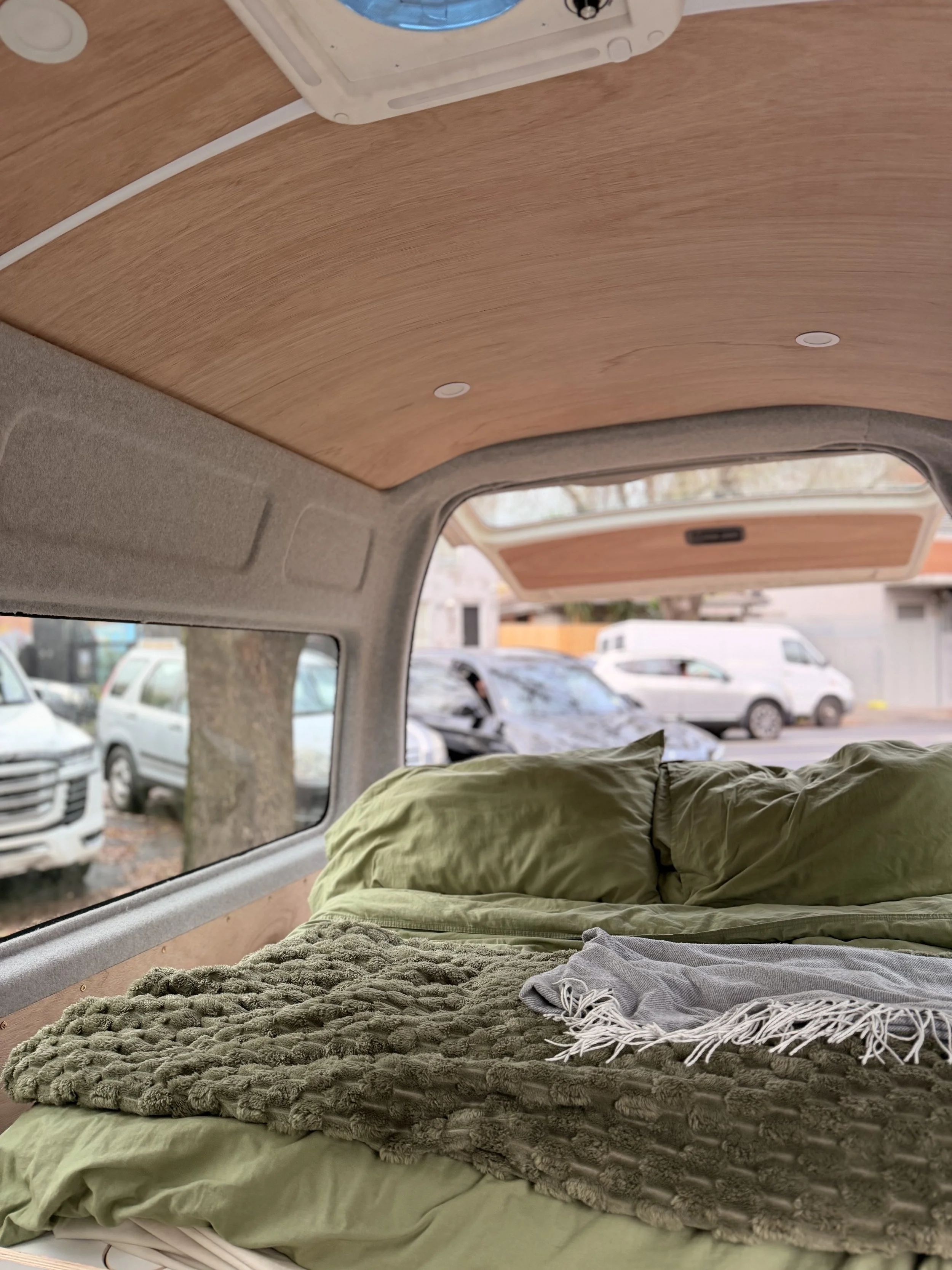inside a camper van with a wooden ceiling, a bed with green and gray bedding, and large windows showing parked cars outside