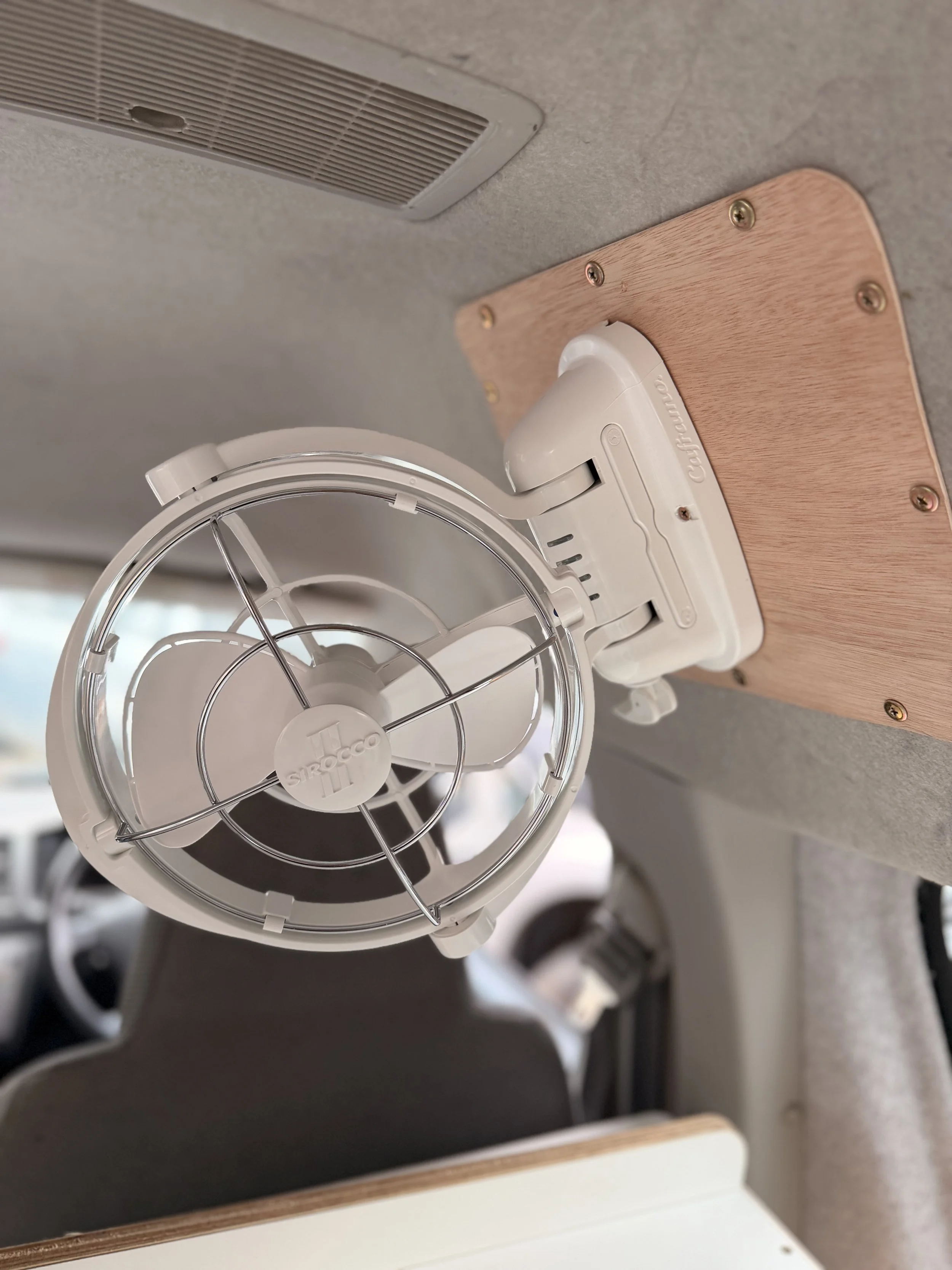 Close-up of a white ventilator or exhaust fan mounted on a wooden board on a gray ceiling, with part of a circular air vent visible below.