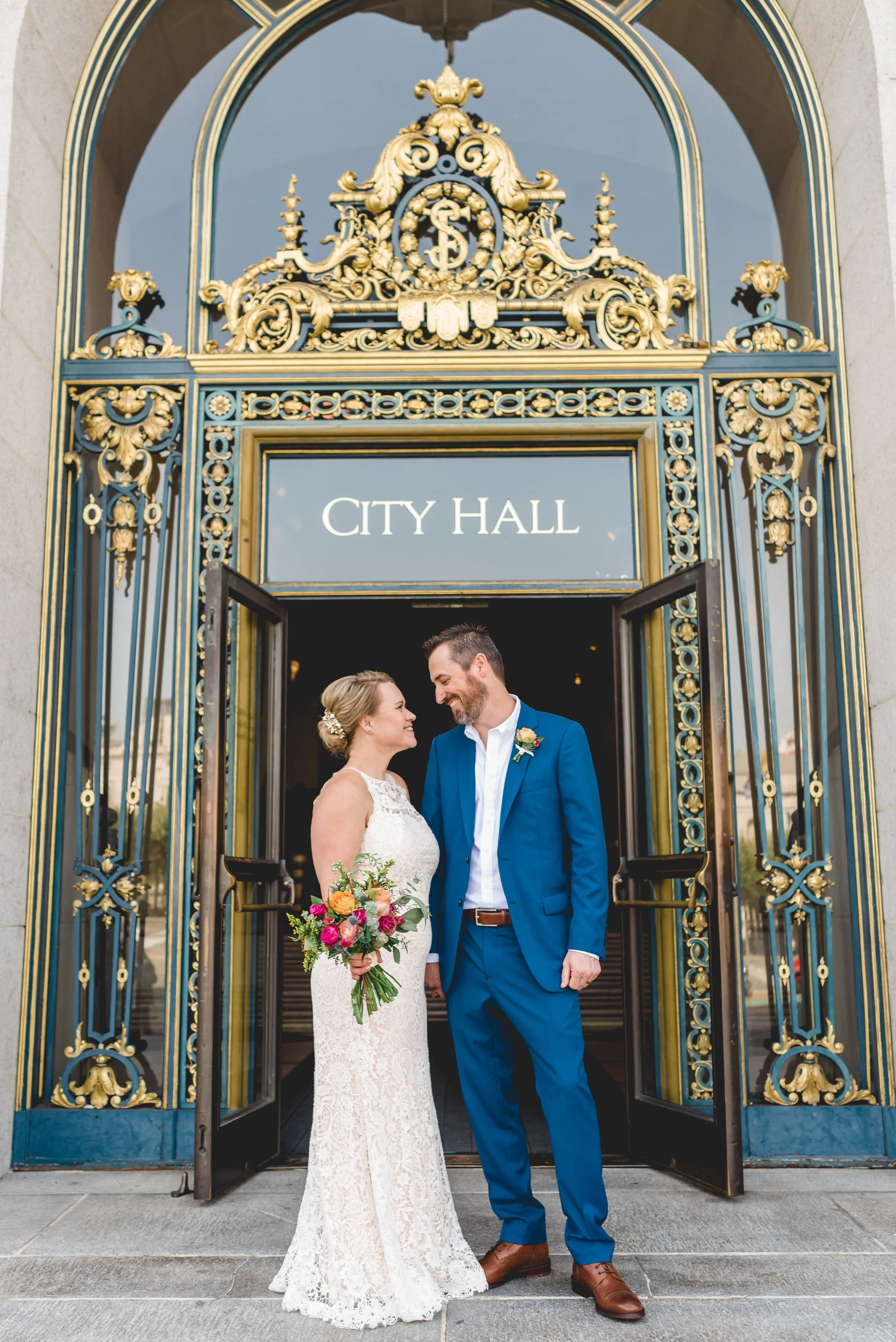 san francisco city hall couple posing in front of ornate doors of city hall with the words city hall above their heads in the window pane