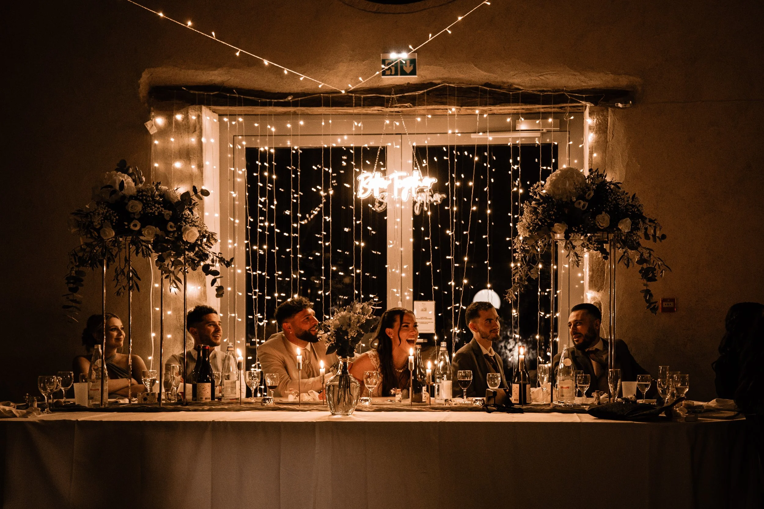 A wedding reception scene showing a bride and five guests seated at a decorated table illuminated by string lights and candles, with floral arrangements and a window with curtains in the background.