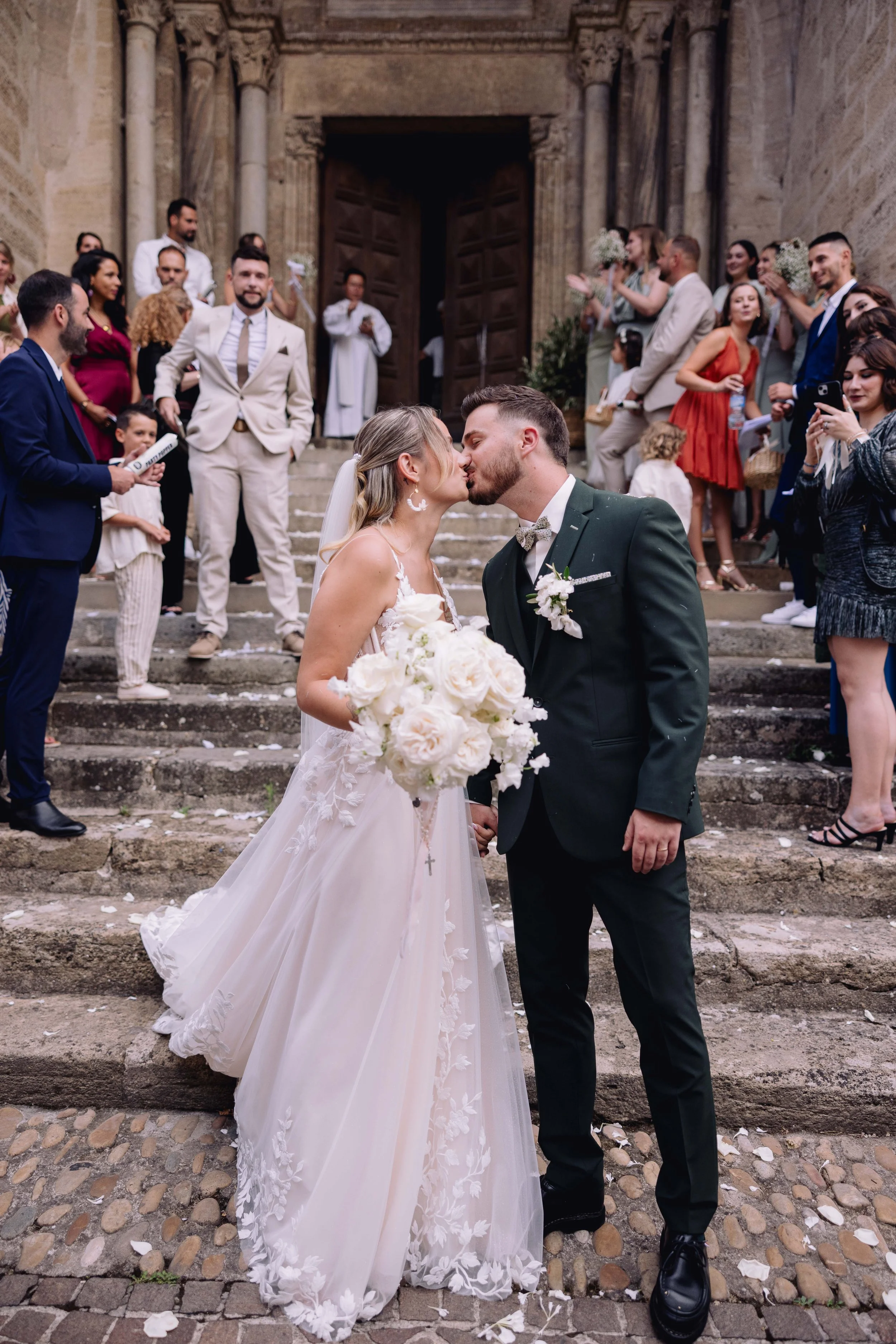 A bride and groom kissing on the steps of a historic building, surrounded by wedding guests, during their wedding celebration.