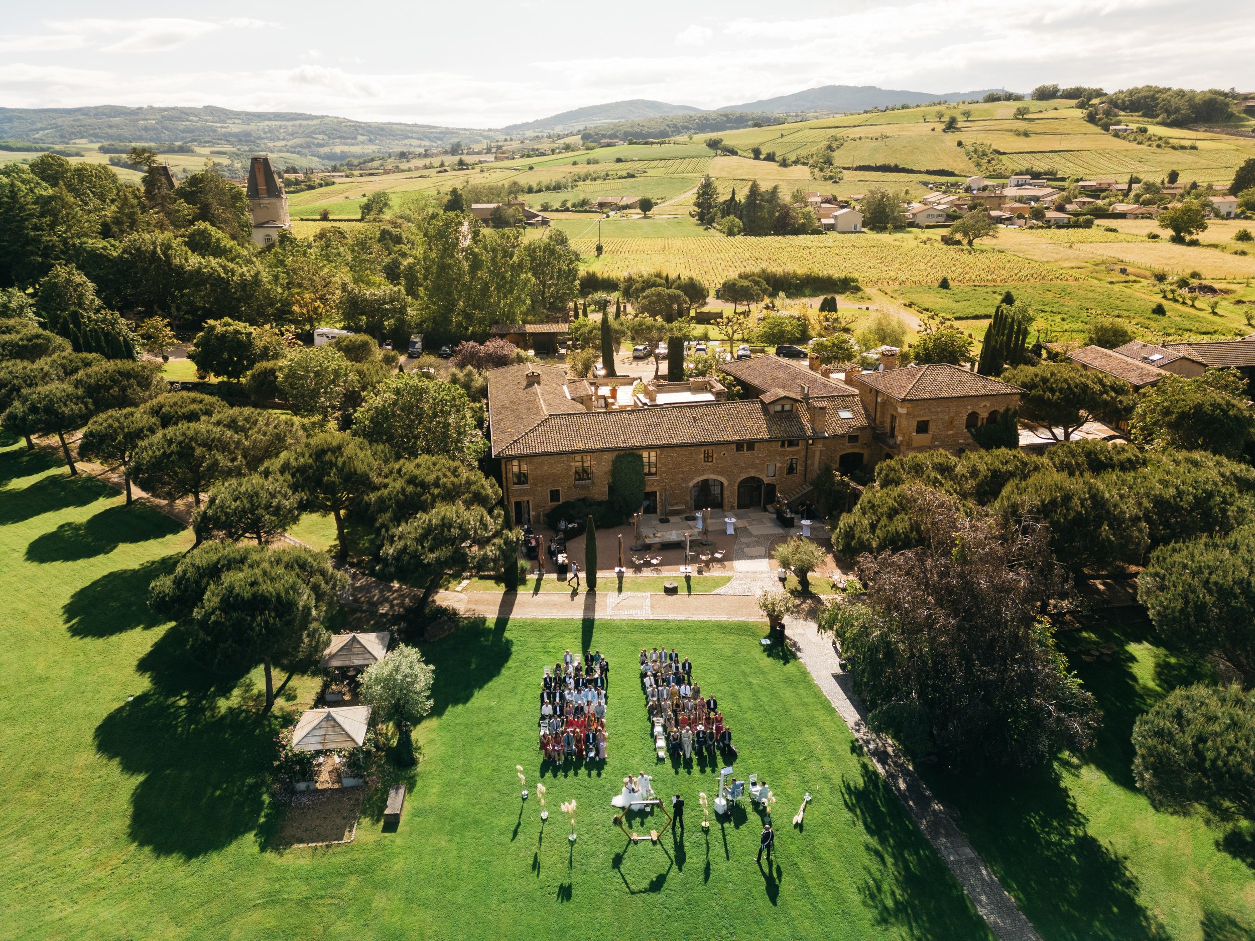 Aerial view of a wedding ceremony outdoors on a lush green lawn surrounded by trees and a large rustic house in a rural landscape with rolling hills and farmland in the background.