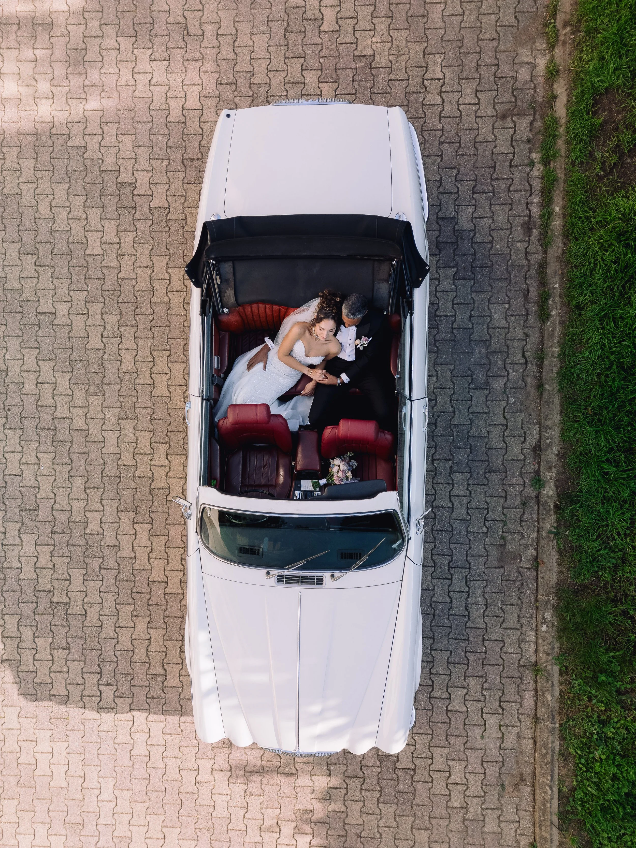 Top-down aerial view of a white vintage convertible car with a bride and groom sitting inside, taken on a brick-paved area.