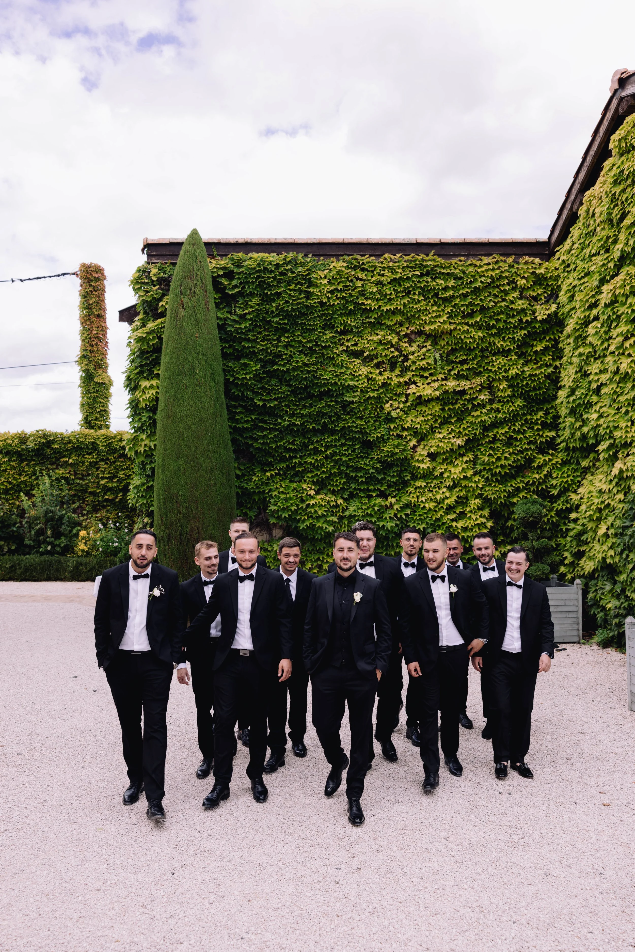 A group of men in tuxedos walking outdoors on a gravel path, with a green ivy-covered wall and tall hedge in the background.