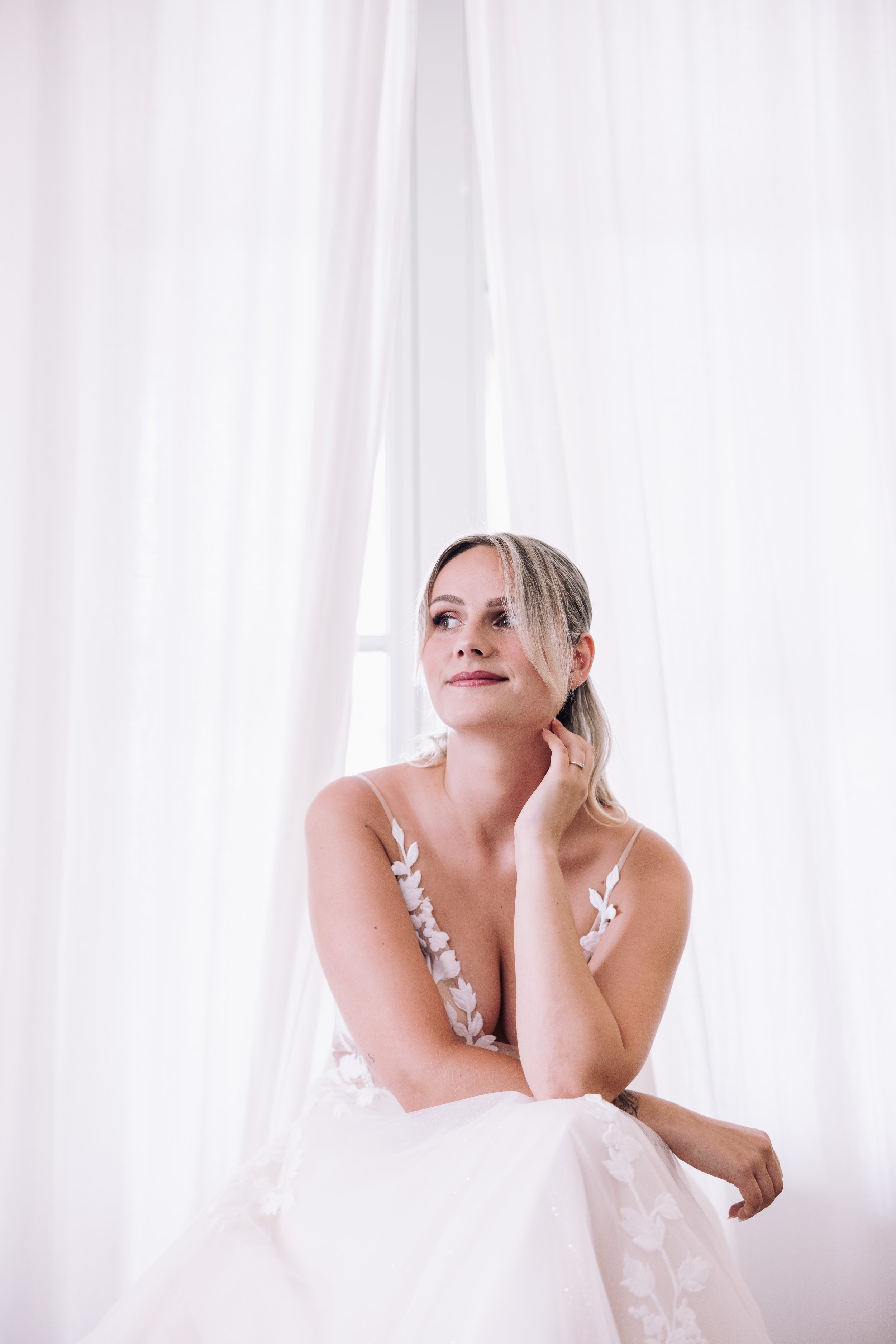 A woman in a wedding dress sitting in front of white curtains, looking thoughtful and posing with her hand on her chin.