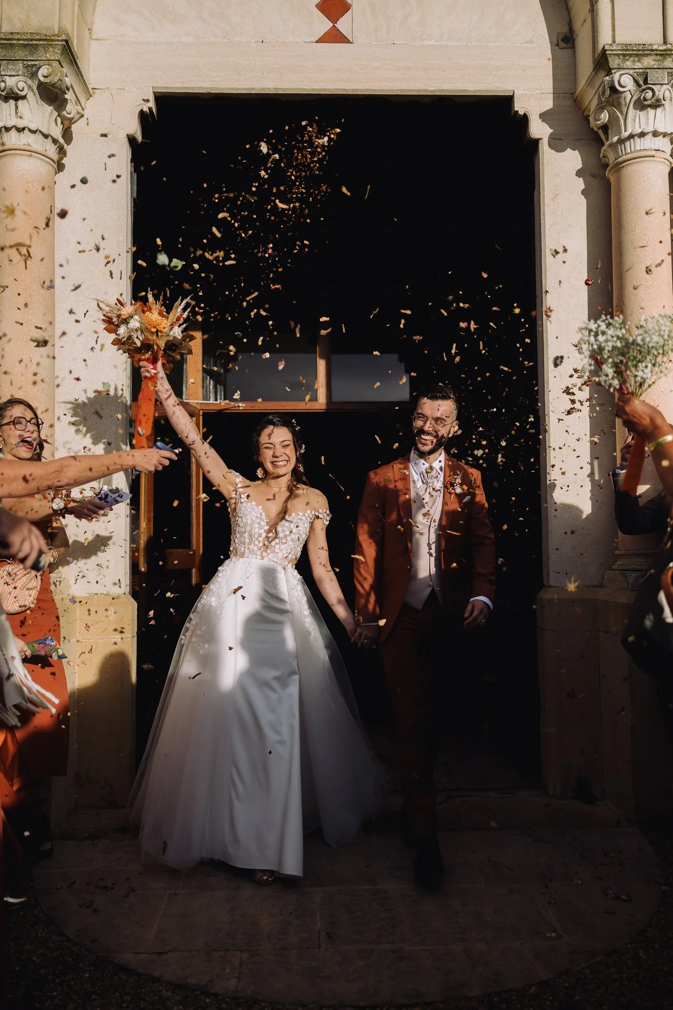 Bride and groom exiting a building, holding hands, celebrating their wedding with guests throwing confetti and flowers in the air.