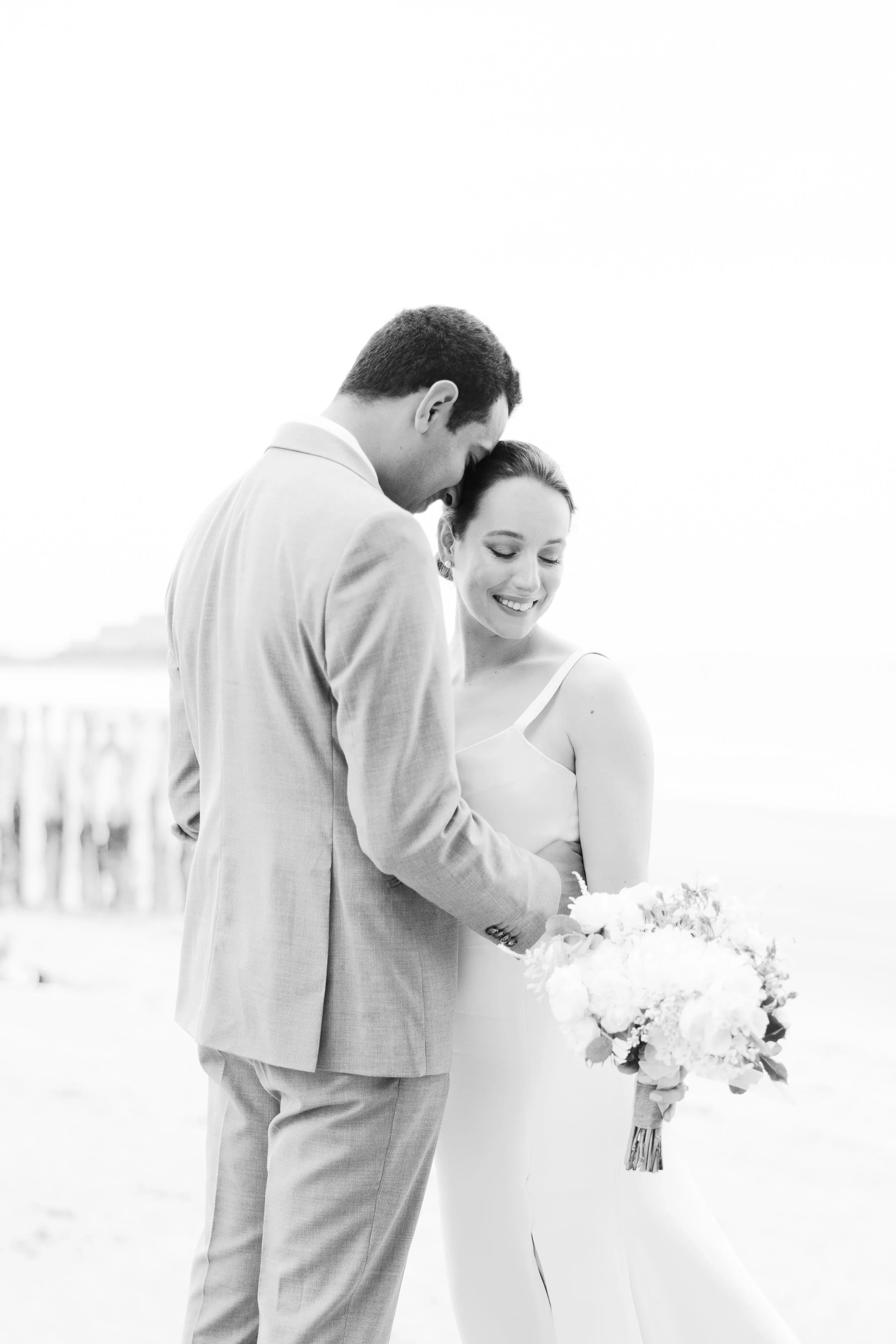 A black and white photograph of a couple embracing at the beach, with a pier in the background, the woman holding a bouquet of flowers.