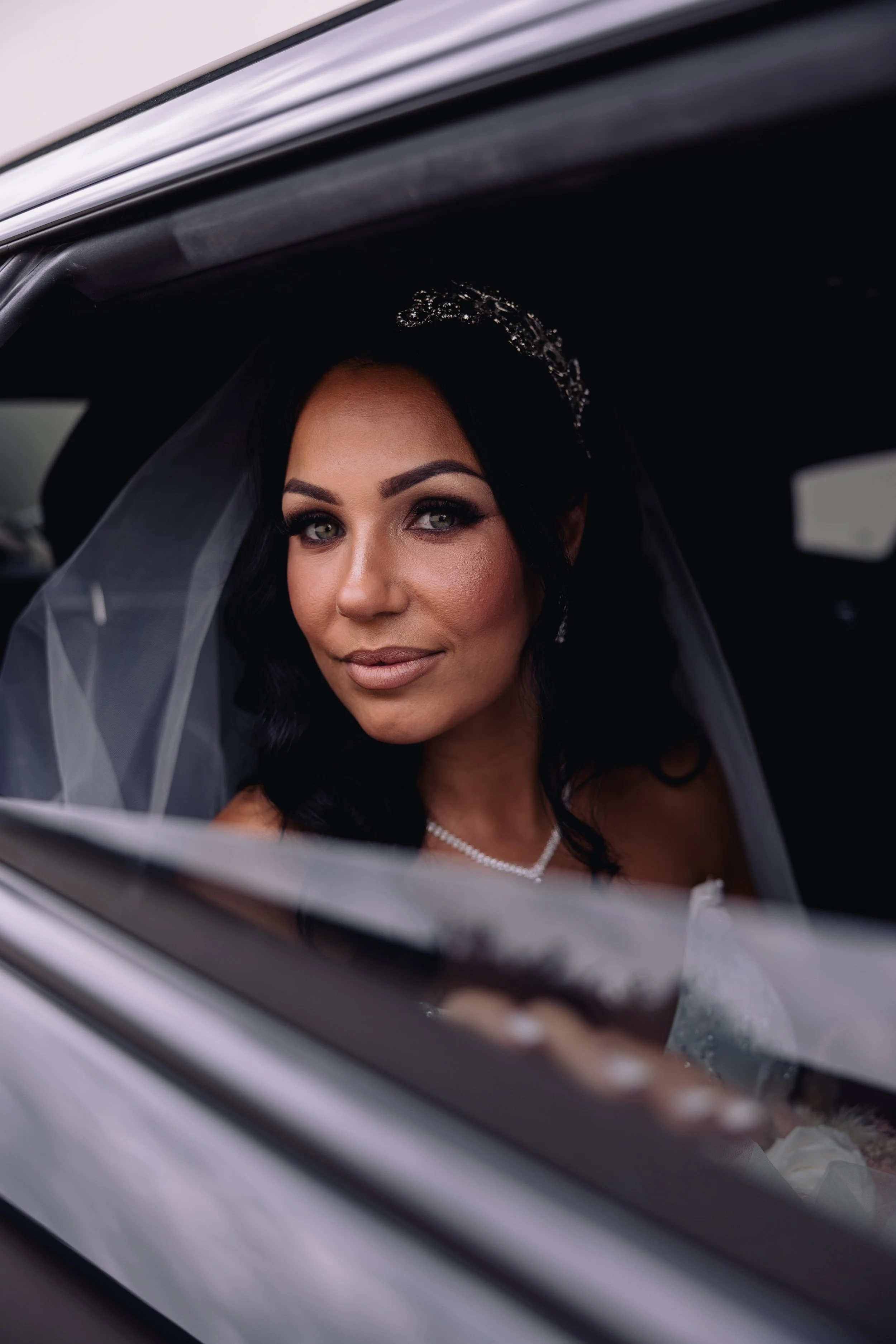 Bride with dark hair, wearing a tiara and pearl necklace, looking out of a car window on her wedding day.
