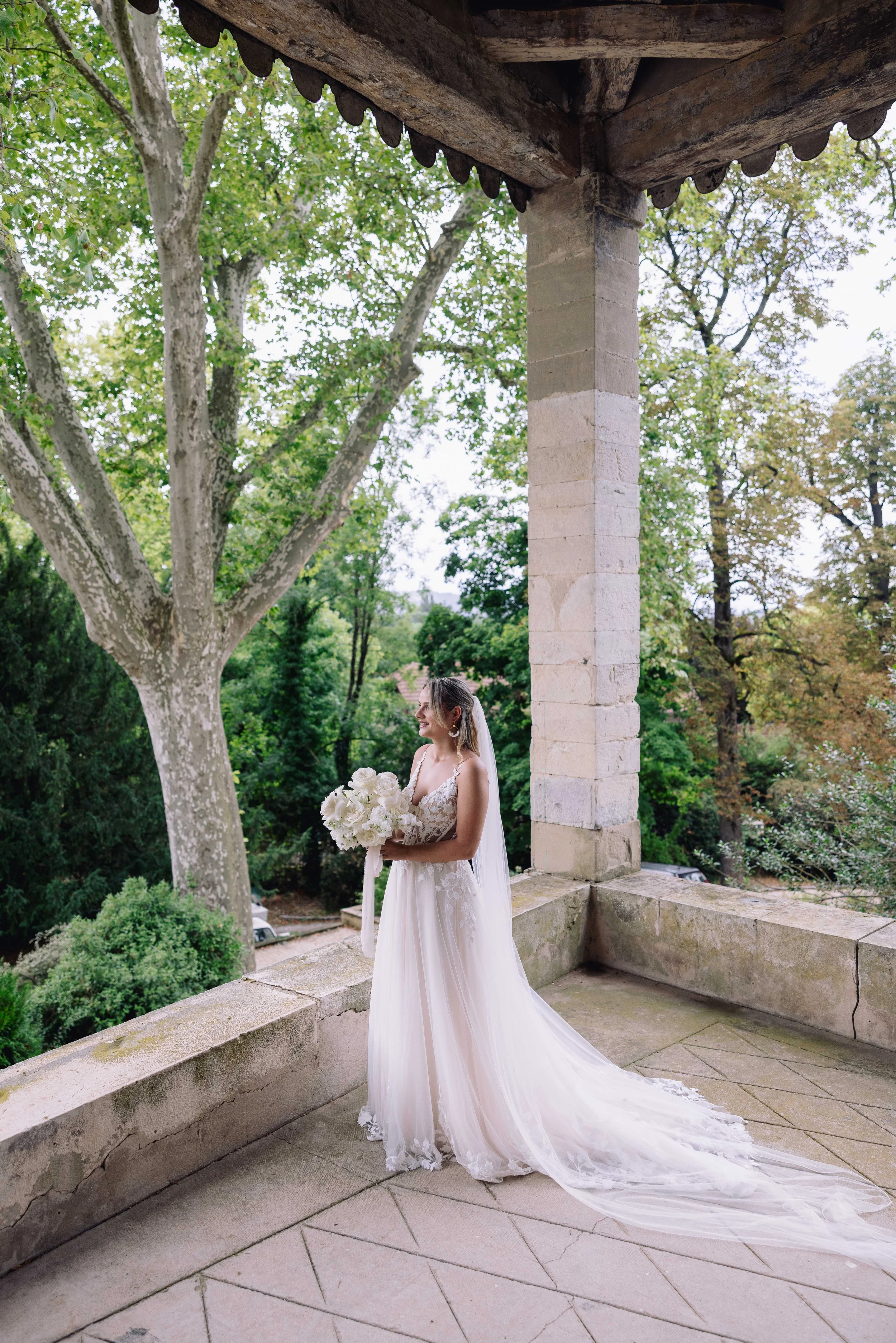 A bride in a wedding dress holding a bouquet of white flowers on a stone balcony with trees in the background.