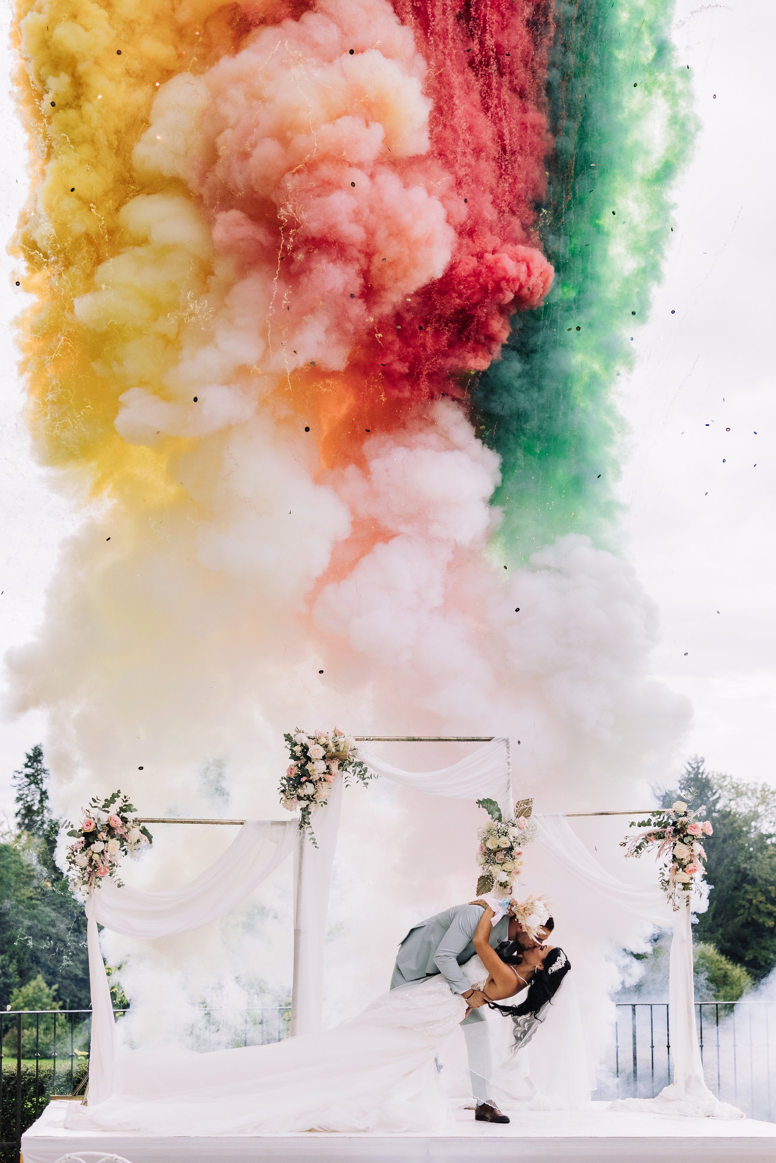 Gay couple kissing at wedding ceremony with colorful smoke explosion overhead.