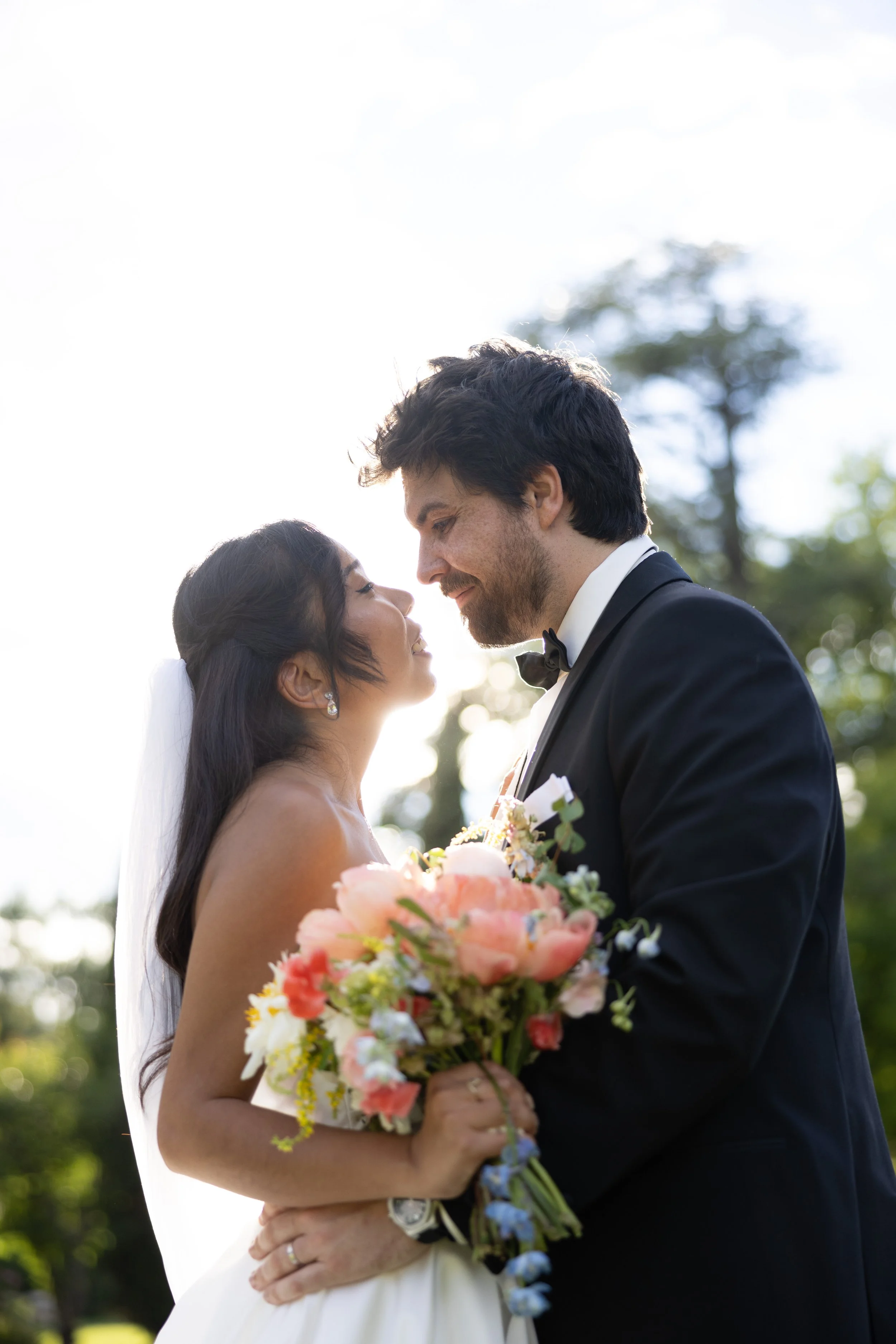 A bride and groom sharing a kiss outdoors, with the bride holding a bouquet of pink, white, and blue flowers, during a wedding ceremony.
