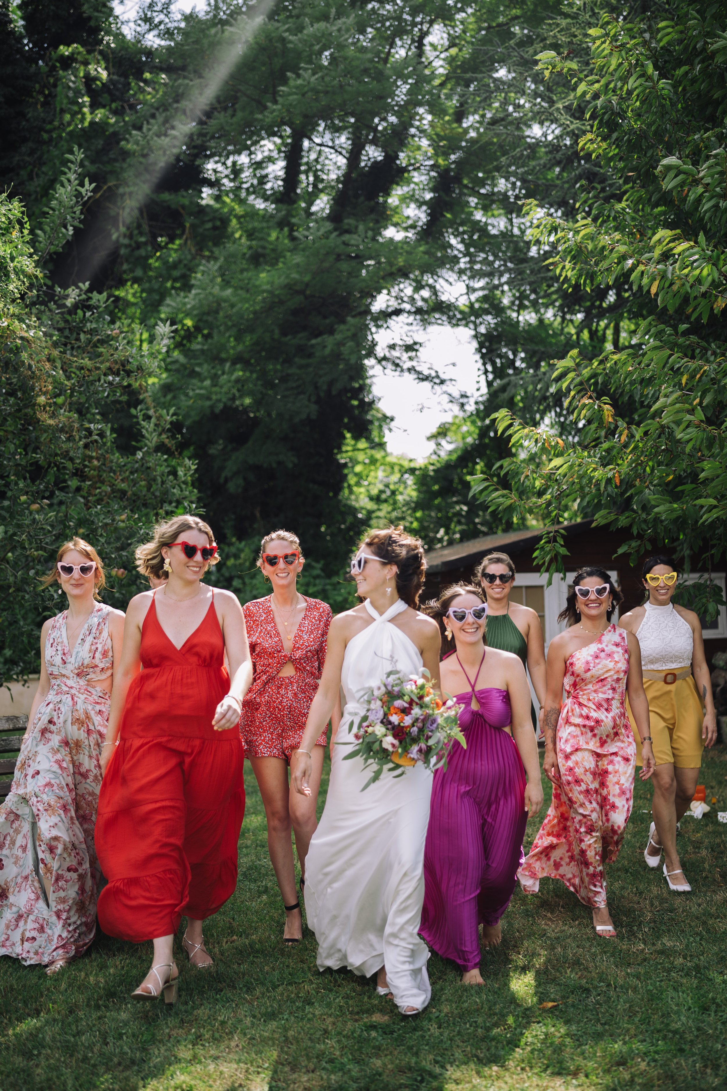A bride in a white dress holding a bouquet walking with a group of women wearing colorful dresses in an outdoor setting surrounded by trees.