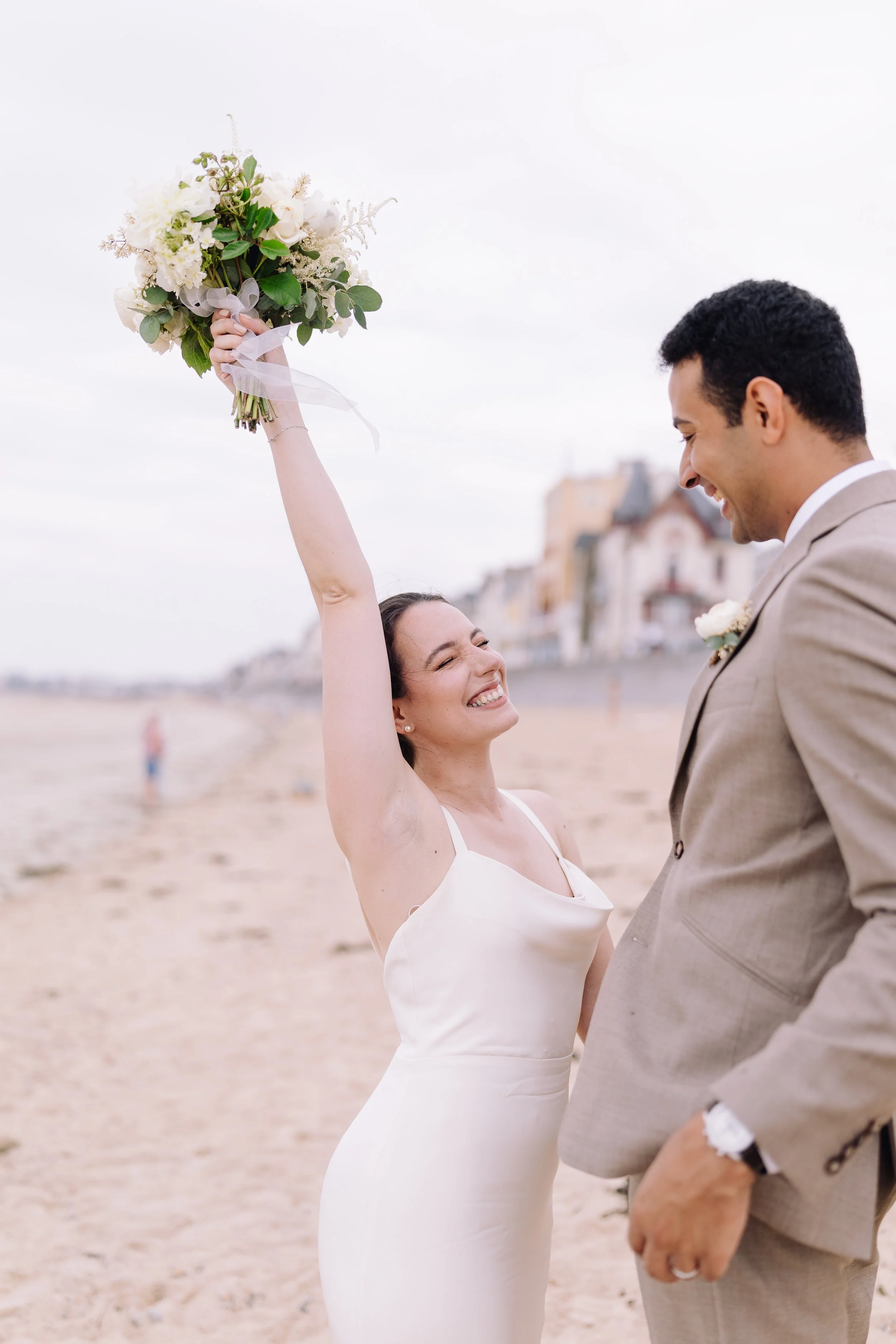 A bride in a white dress holding a bouquet and smiling as she celebrates with her groom on the beach.