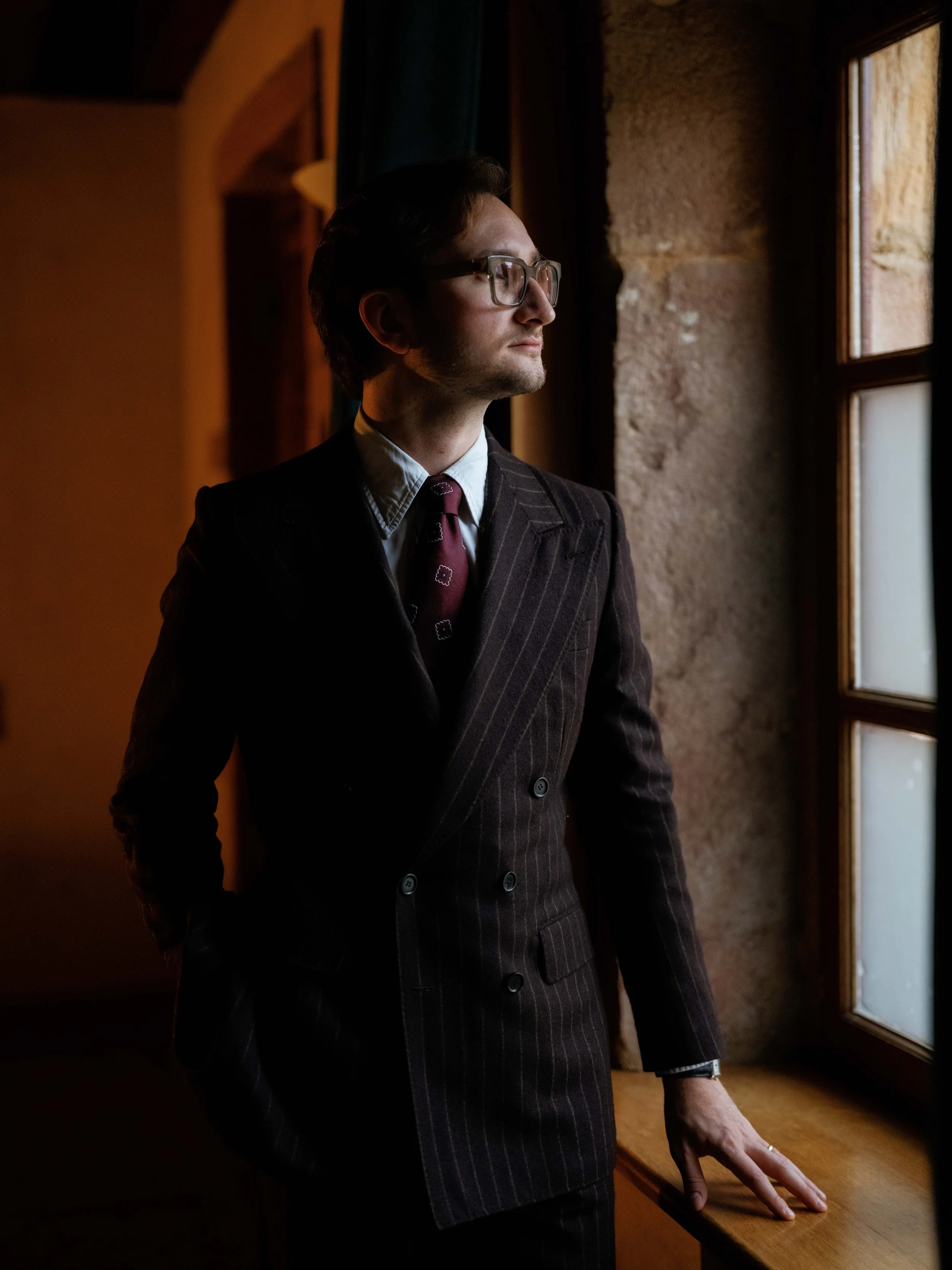 A man in a dark pinstripe suit with a white shirt and patterned tie, wearing glasses, stands by a window looking outside, with one hand resting on a wooden windowsill.
