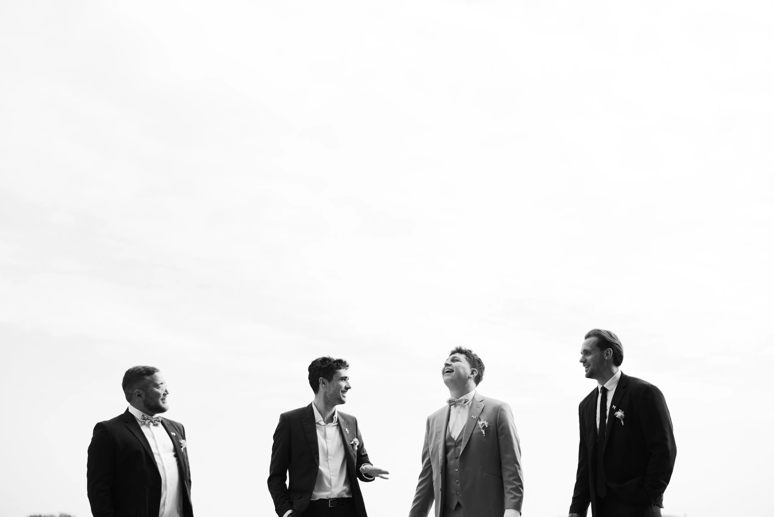Four men dressed in suits standing outdoors, engaging in conversation and smiling, with a cloudy sky in the background.