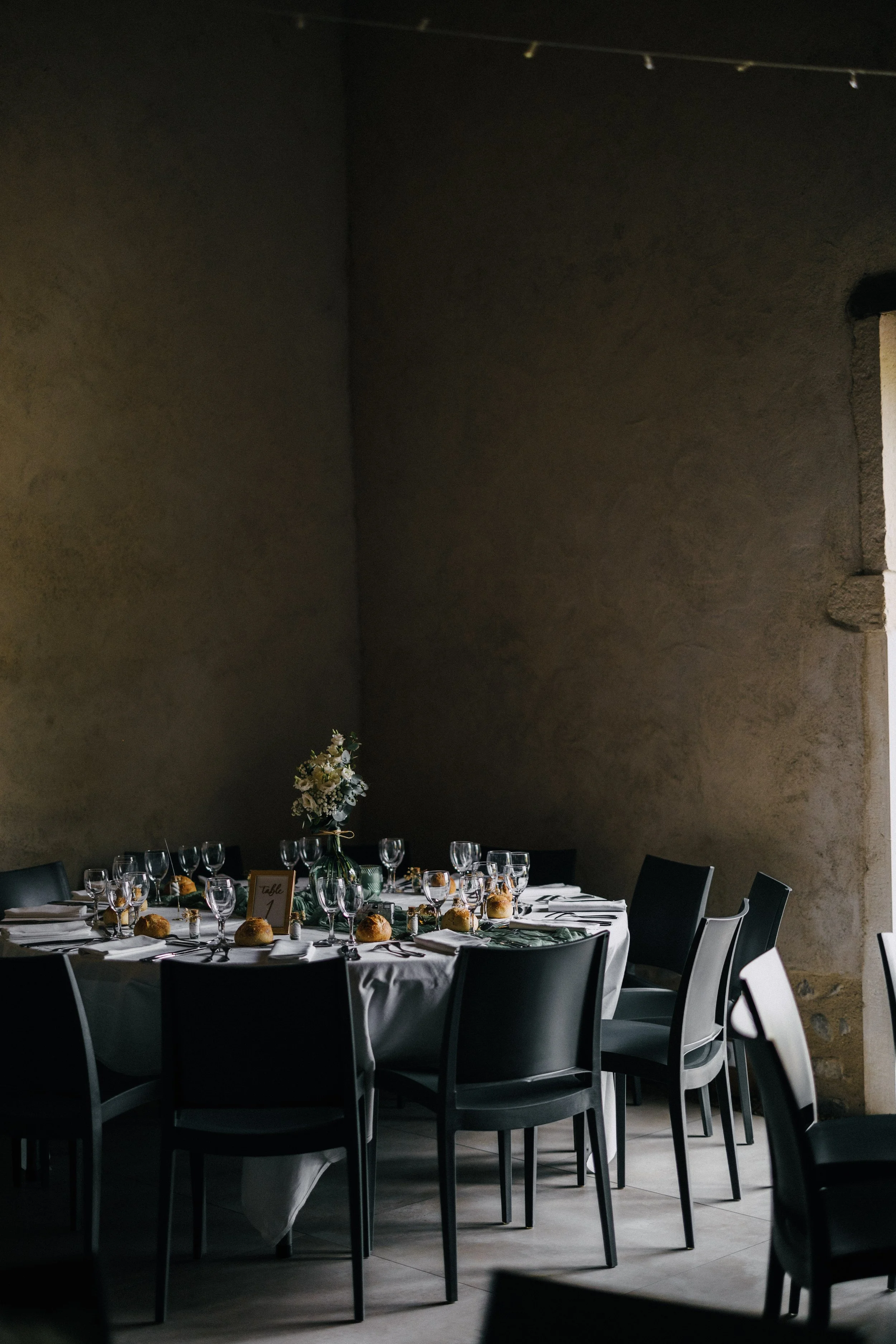 A round banquet table set for an event with black chairs, a white tablecloth, a floral centerpiece in a glass vase, wine glasses, plates, silverware, napkins, and bread rolls. The setting is inside a room with dark, textured walls and natural light c