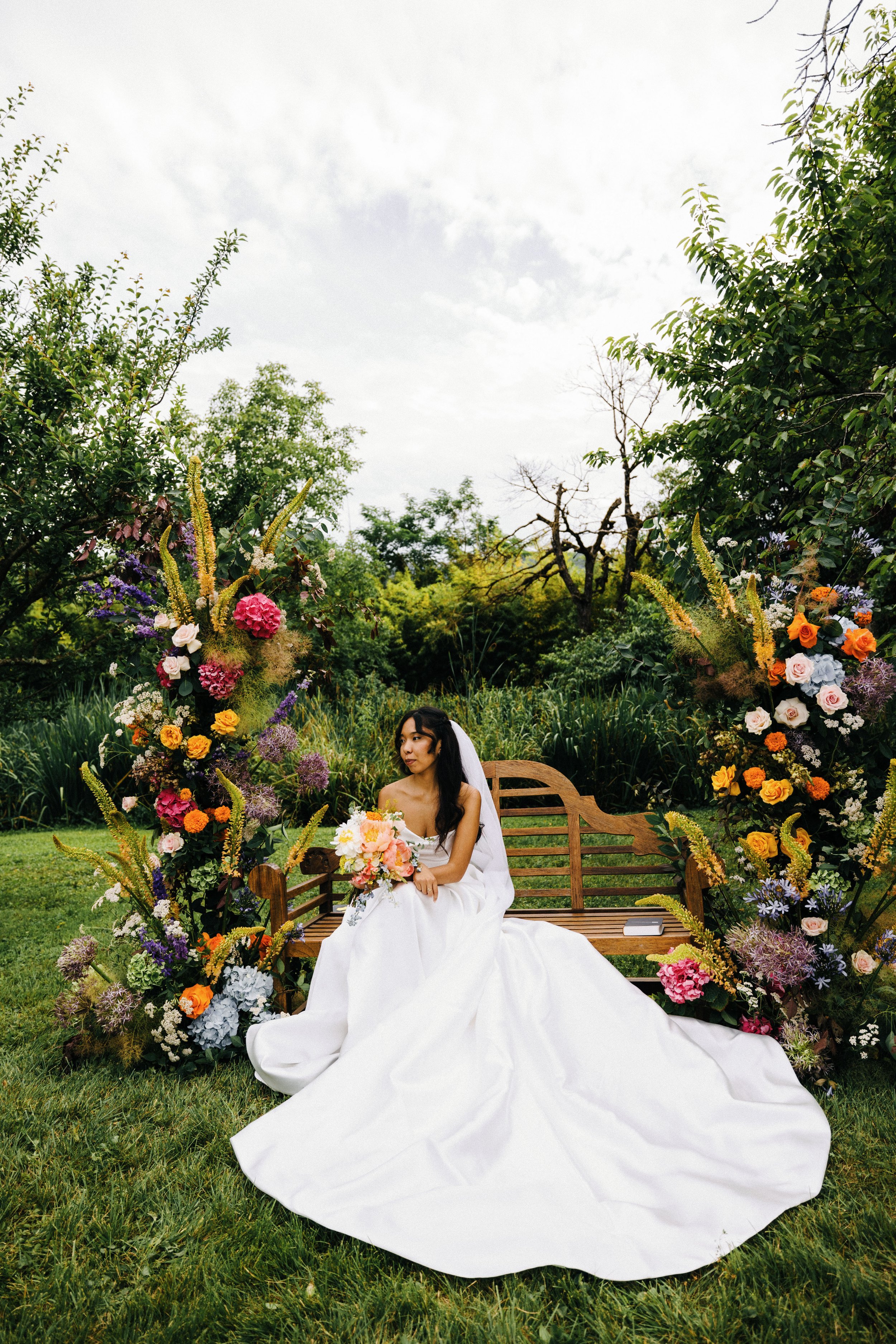 A bride in a white wedding dress sitting on a wooden bench outdoors, surrounded by colorful flower arrangements and greenery.