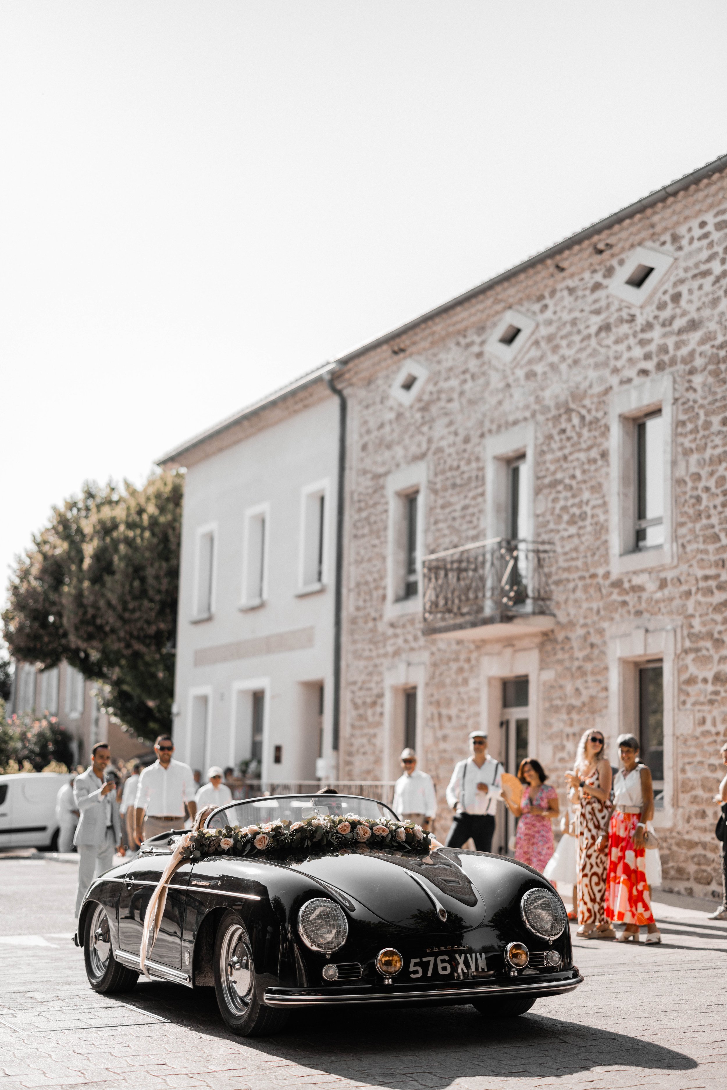 Black vintage convertible car decorated with flowers and ribbons is parked on a cobblestone street. People dressed in semi-formal and colorful clothes are gathered nearby on a sunny day, in front of stone and white buildings.