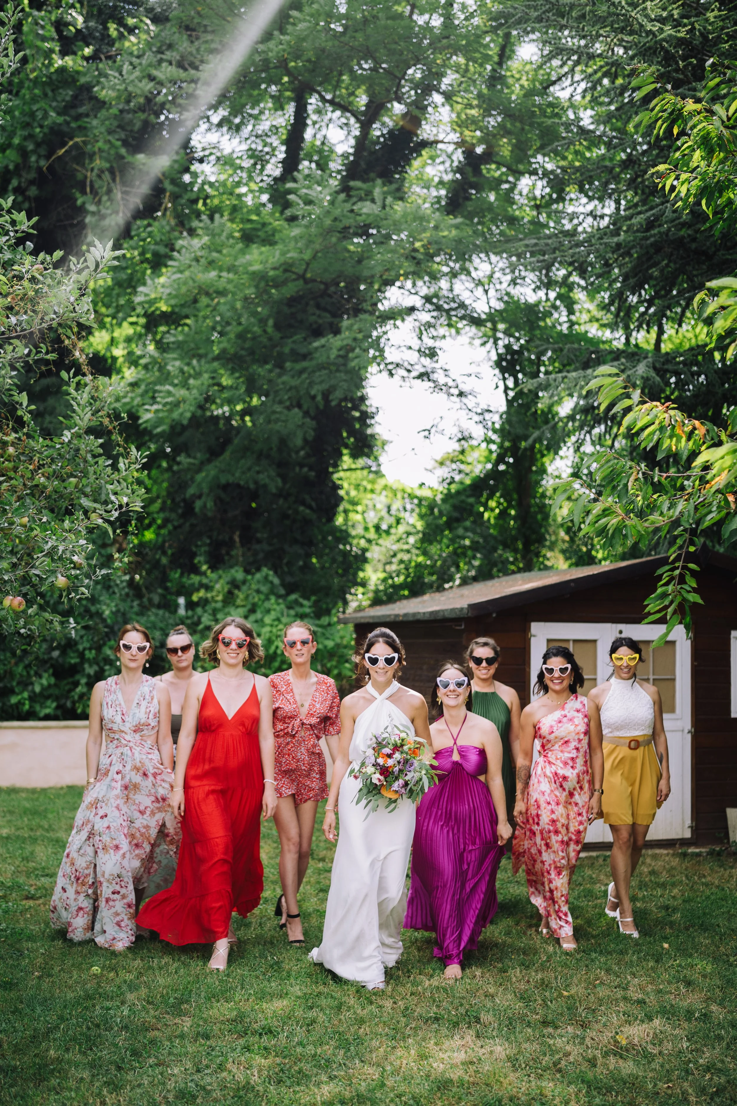 A group of women walking outdoors on grass, dressed in colorful dresses, wearing sunglasses, with a wooden shed and trees in the background. One woman is holding a bouquet of flowers.