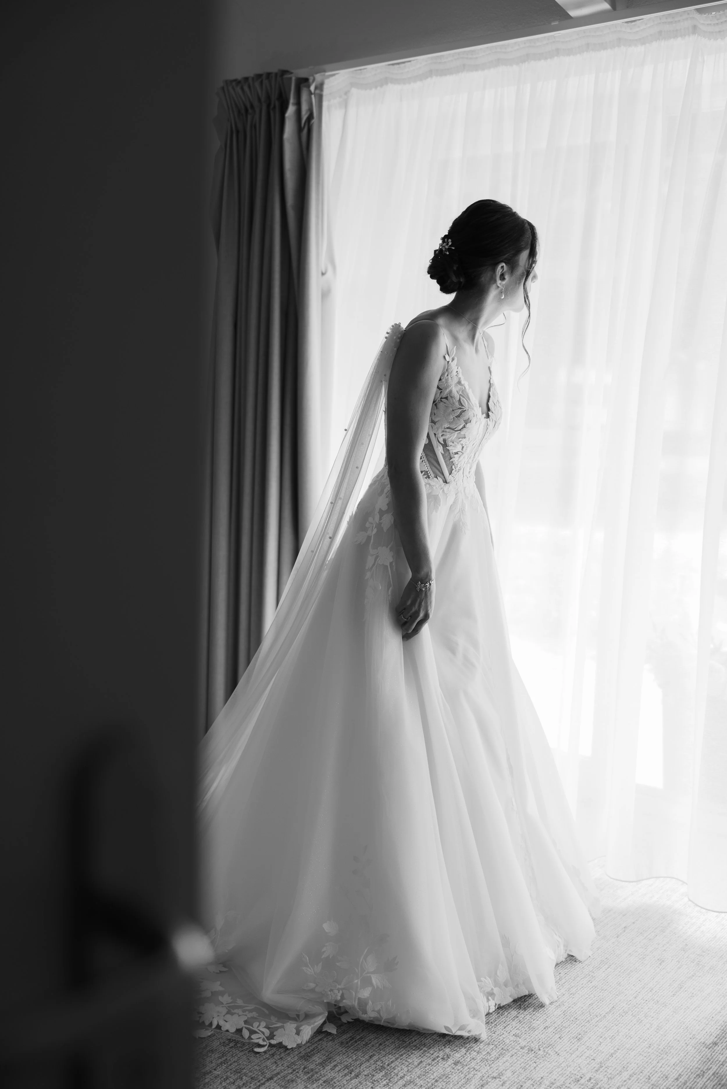 A bride in a wedding gown gazes out a window, with curtains and sheer drapes, in a black-and-white photograph.