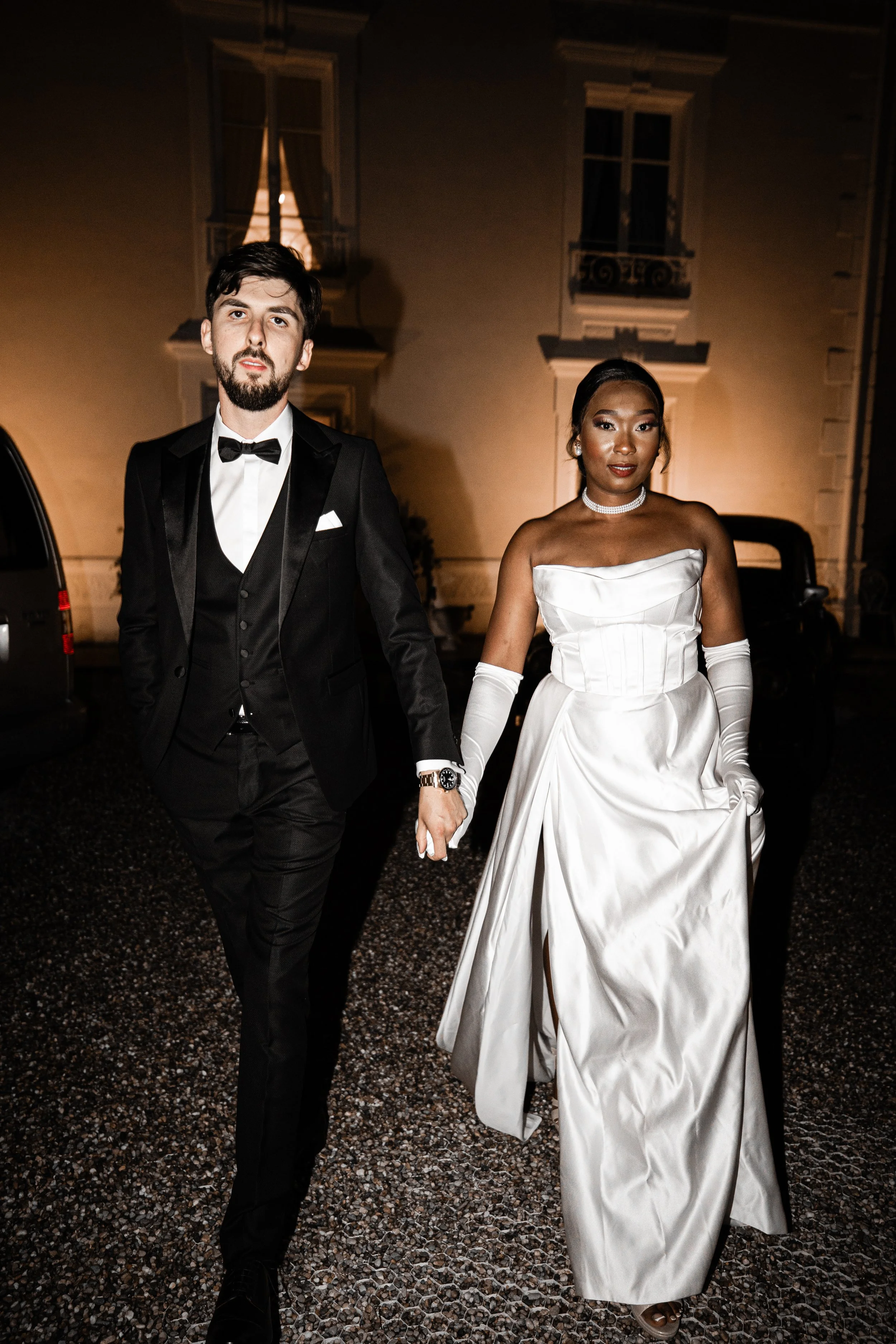 A couple dressed in elegant formal attire, holding hands, walking outdoors at night, with a building featuring large windows in the background.