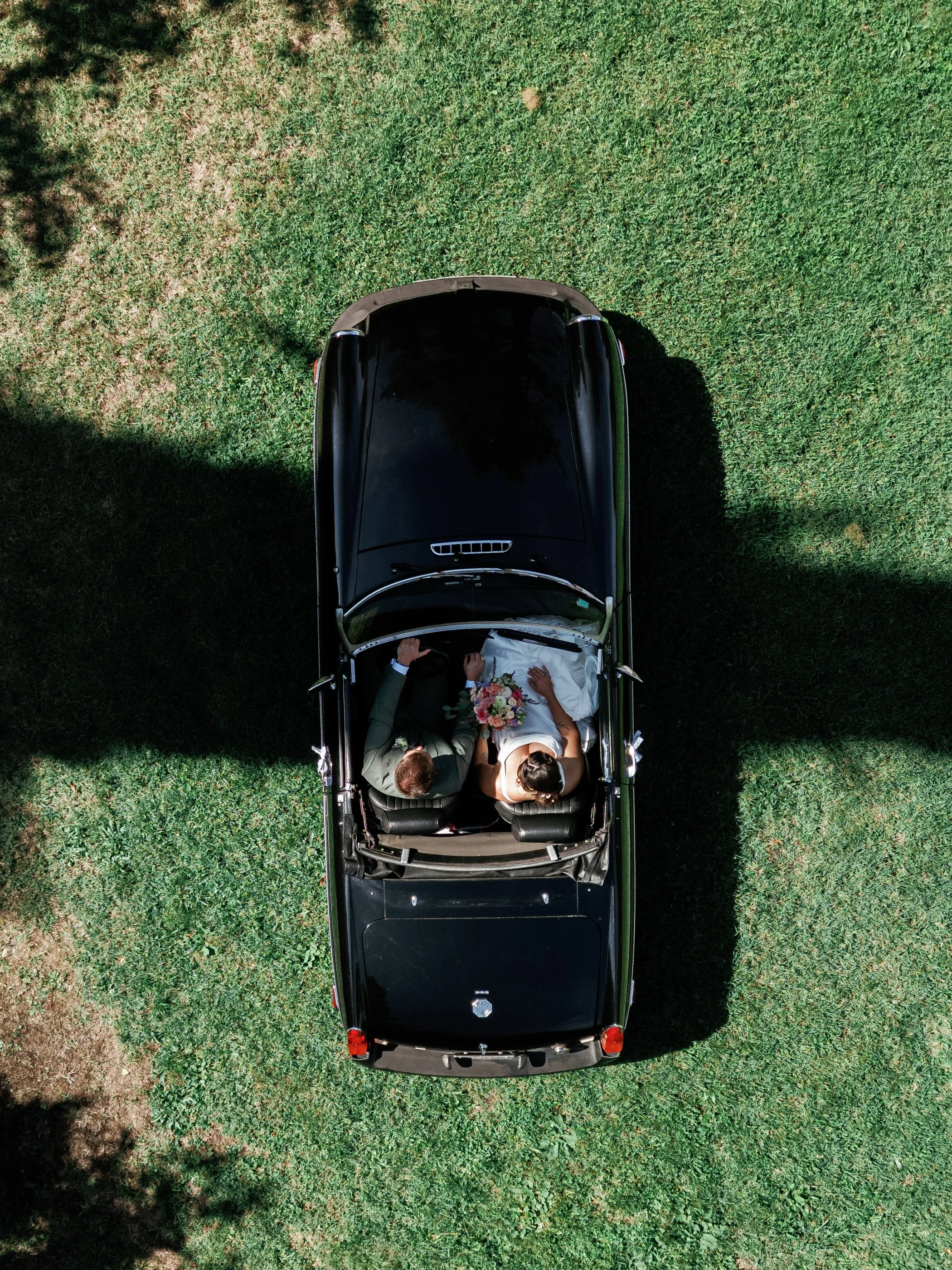 A wedding couple lying on the front seats of a black convertible car, taken from above on a grassy area with shadows of trees.