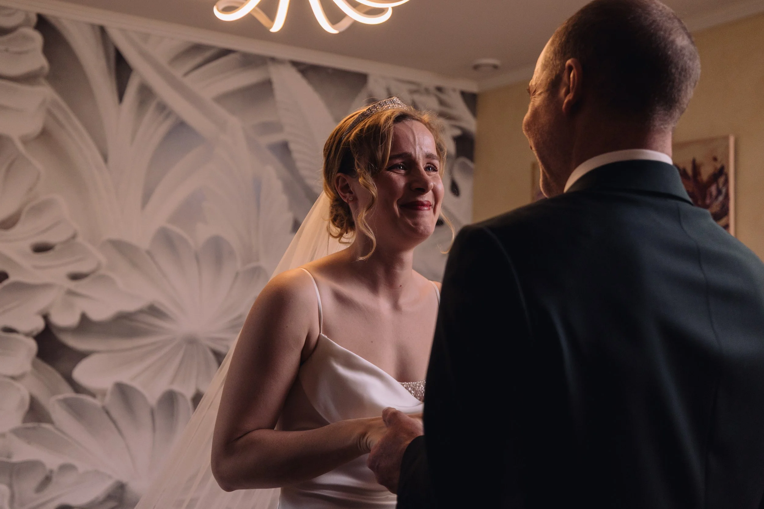 A bride and groom share an emotional moment during their wedding vows indoors, with the bride visibly tearful and the groom holding her hand.