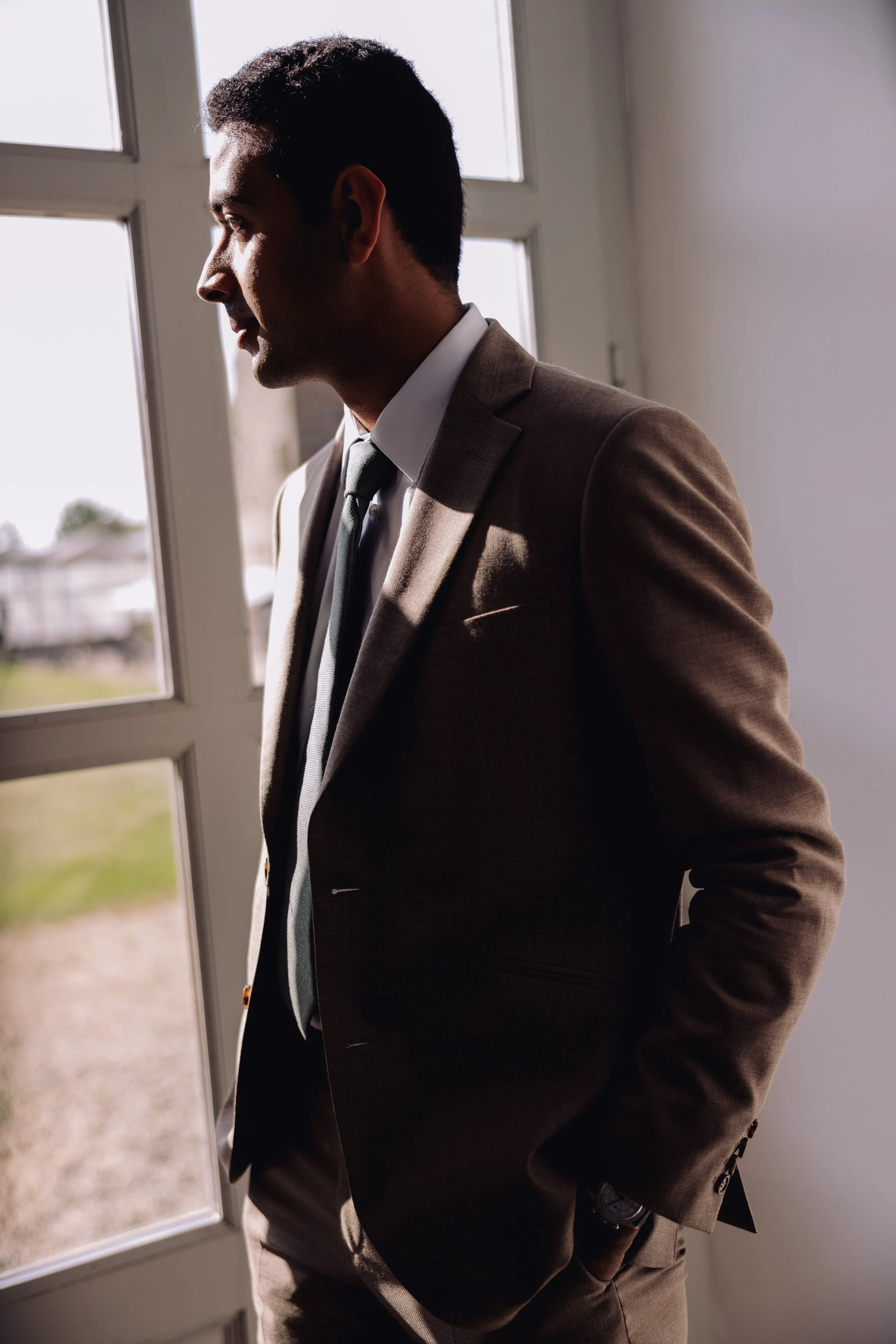 Man in a brown suit and tie standing near a large window with sunlight, looking outside, with hands in pockets.