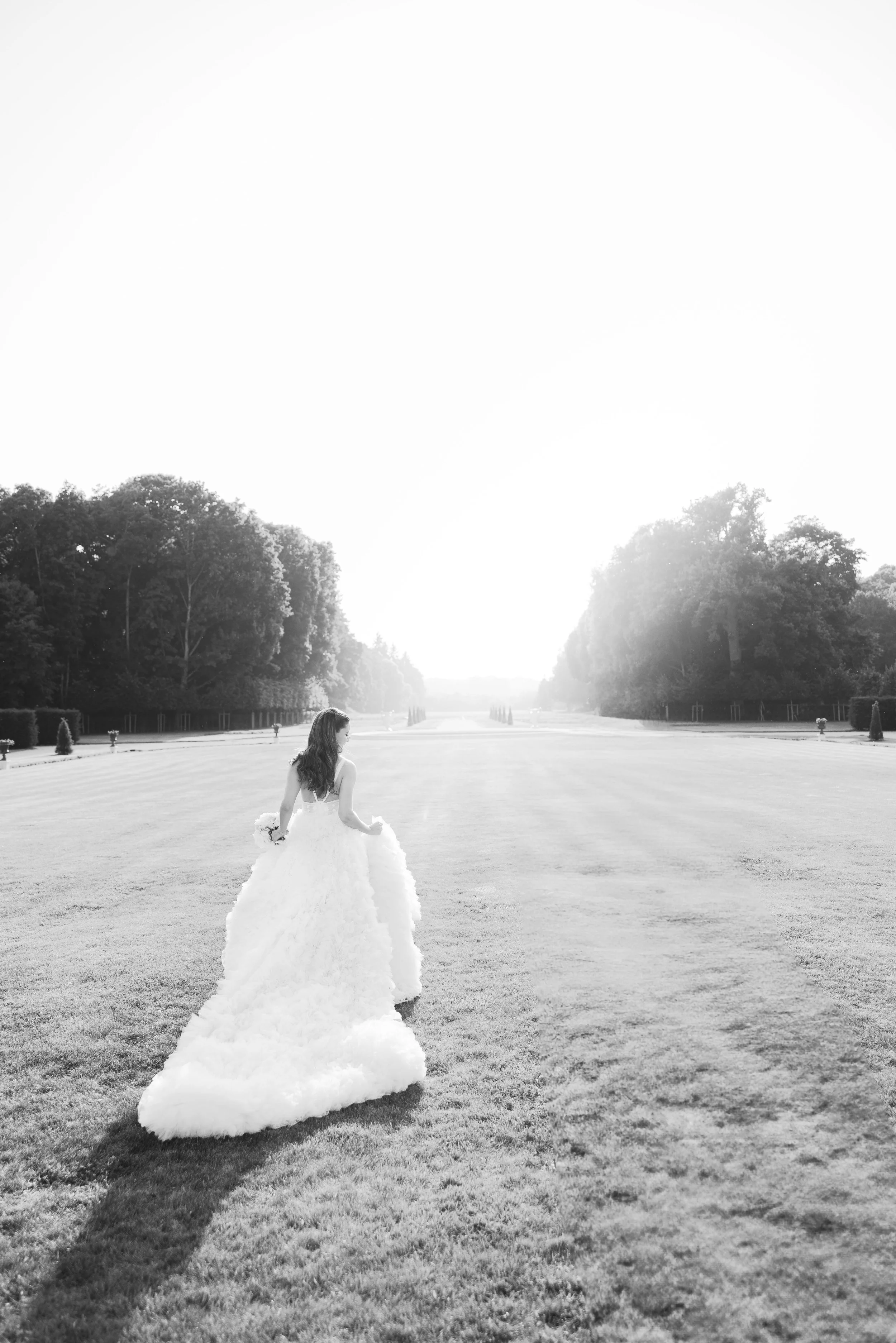 A woman in a wedding dress walking across an open field with trees on either side, taken in black and white.