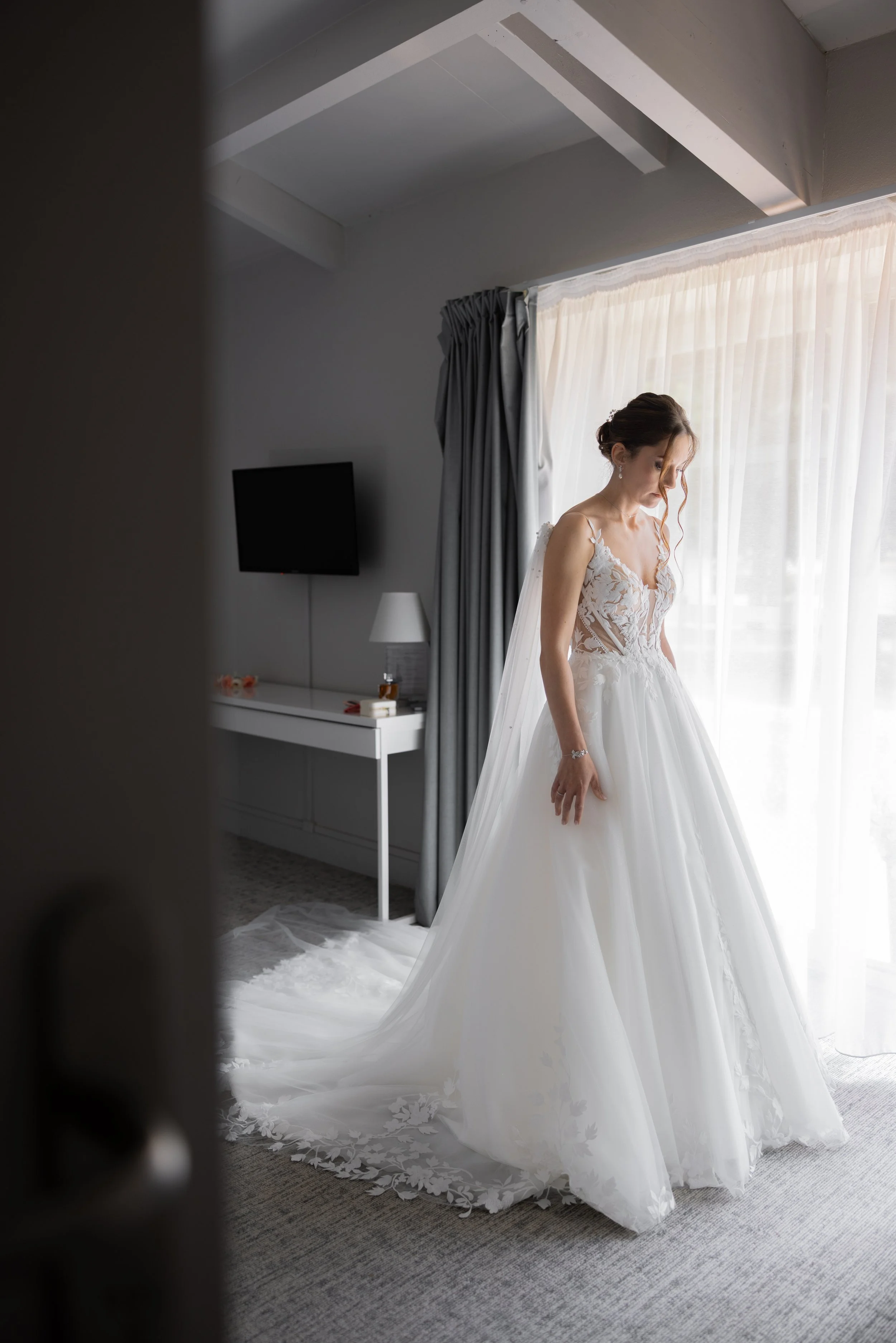 A bride in a white wedding gown standing by a window, looking down, with natural light illuminating her.