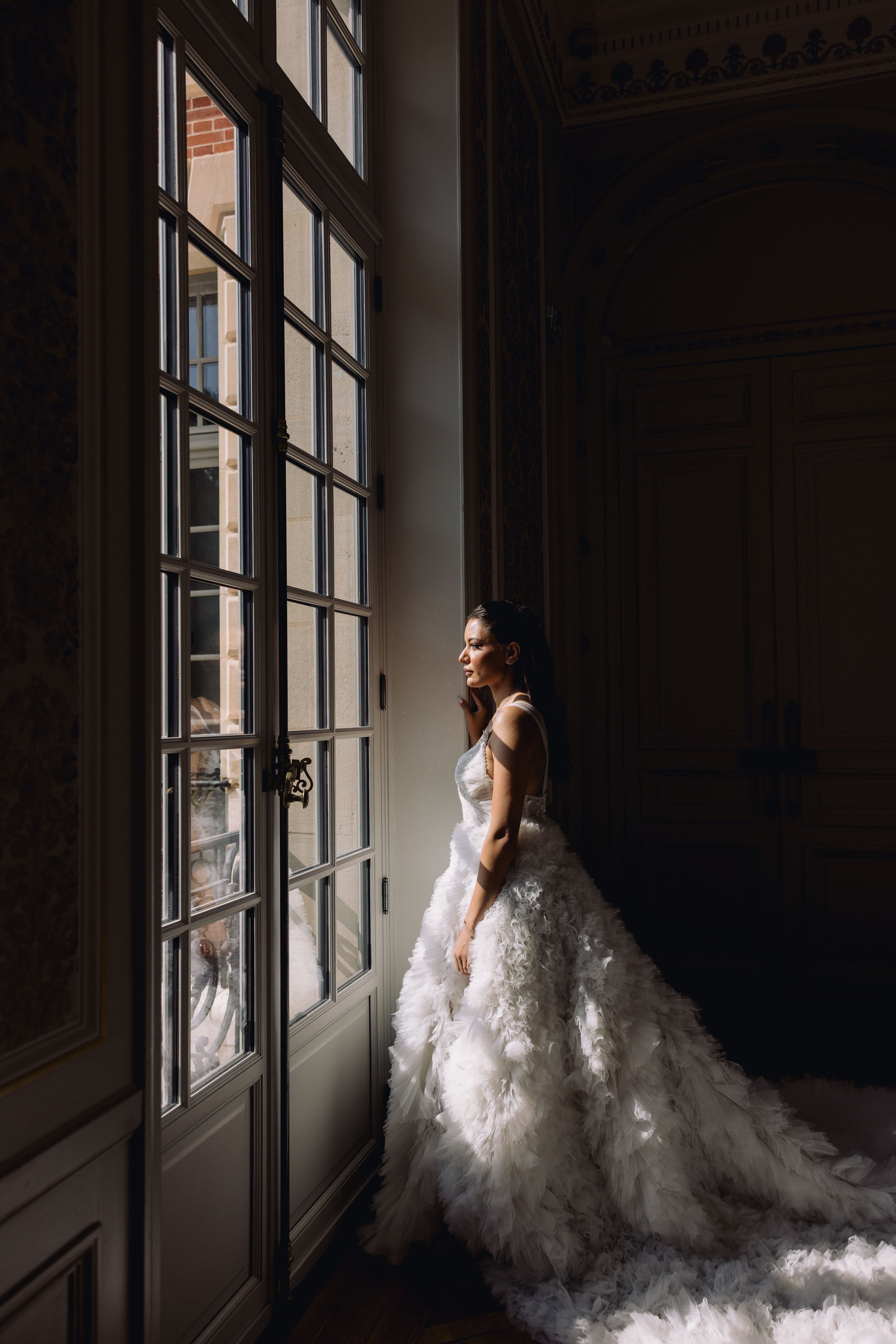 A woman in a white wedding gown standing by a large window, looking outside, with sunlight illuminating her face and dress inside a dimly lit room.