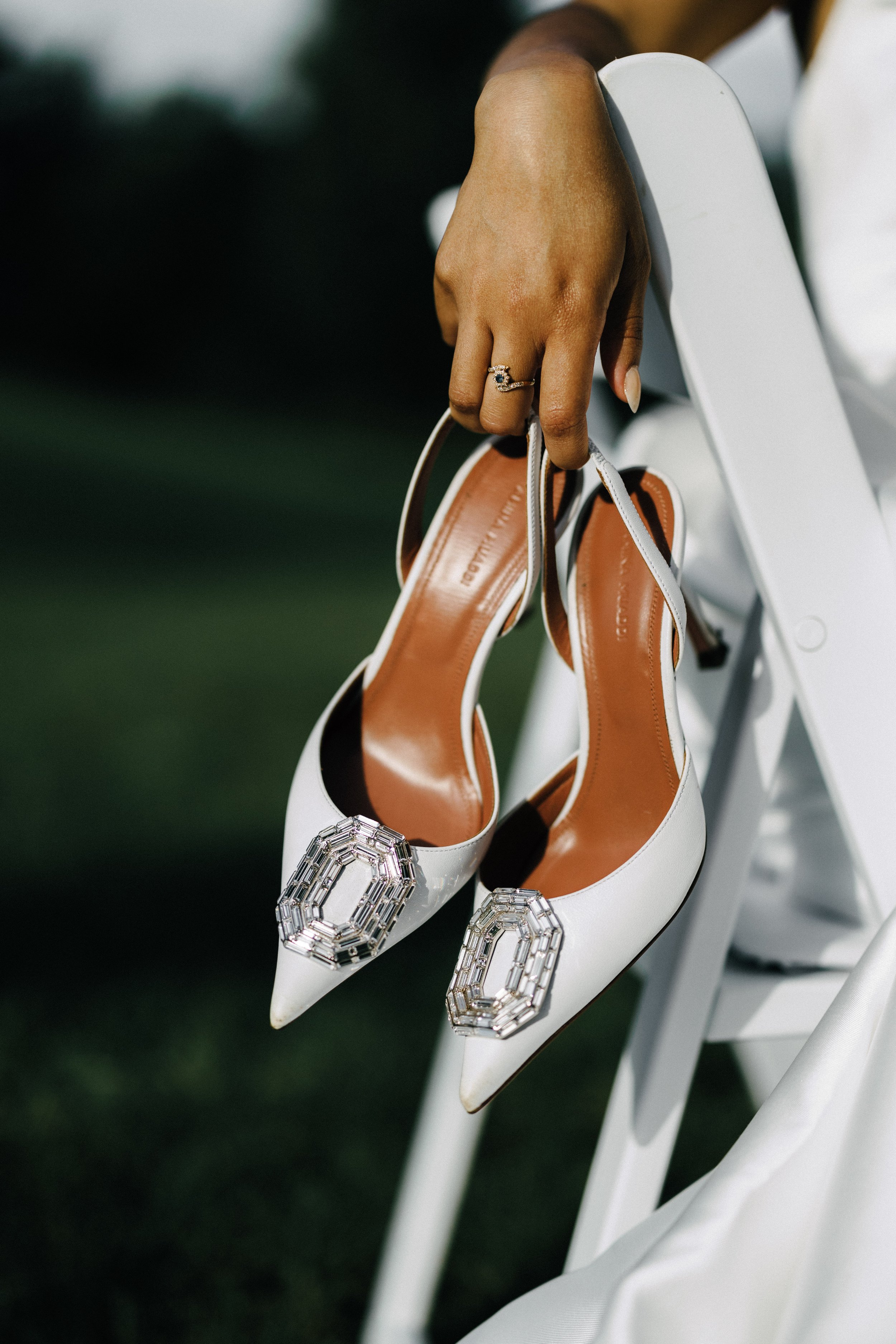 A person's hand with a ring is holding white high-heeled shoes with decorative buckle embellishments, hanging over the edge of a white chair.