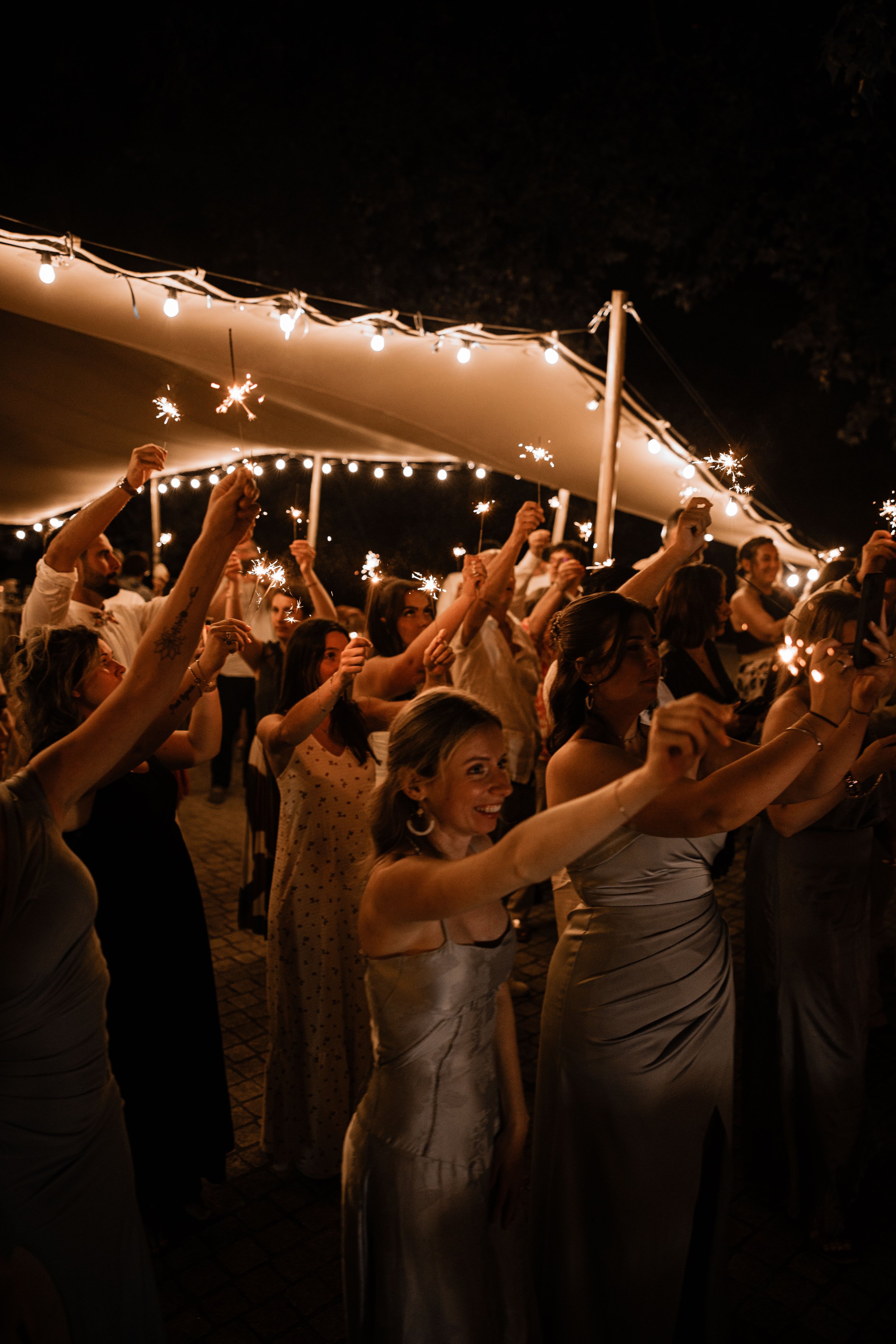People celebrating at night holding sparklers under string lights at an outdoor event.