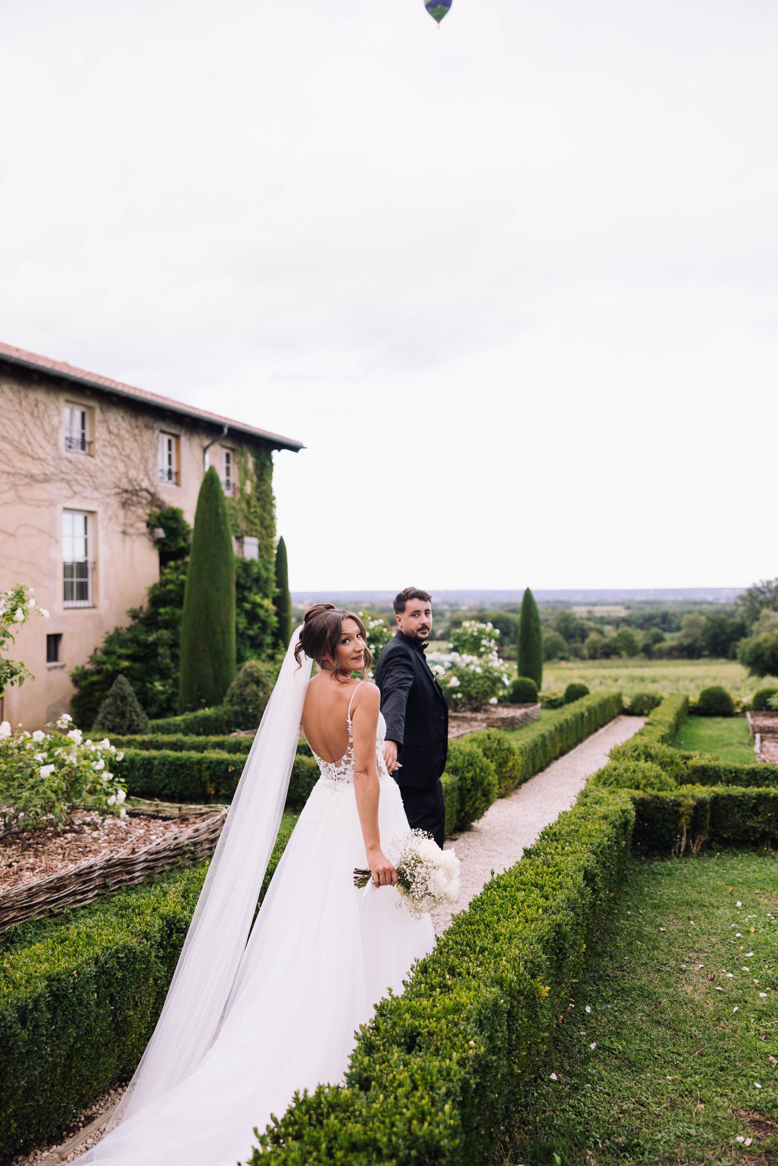 Bride in a wedding dress holding a bouquet turned to look back, groom in a black suit, standing on a garden path bordered by trimmed bushes, with a stone building and trees in the background.