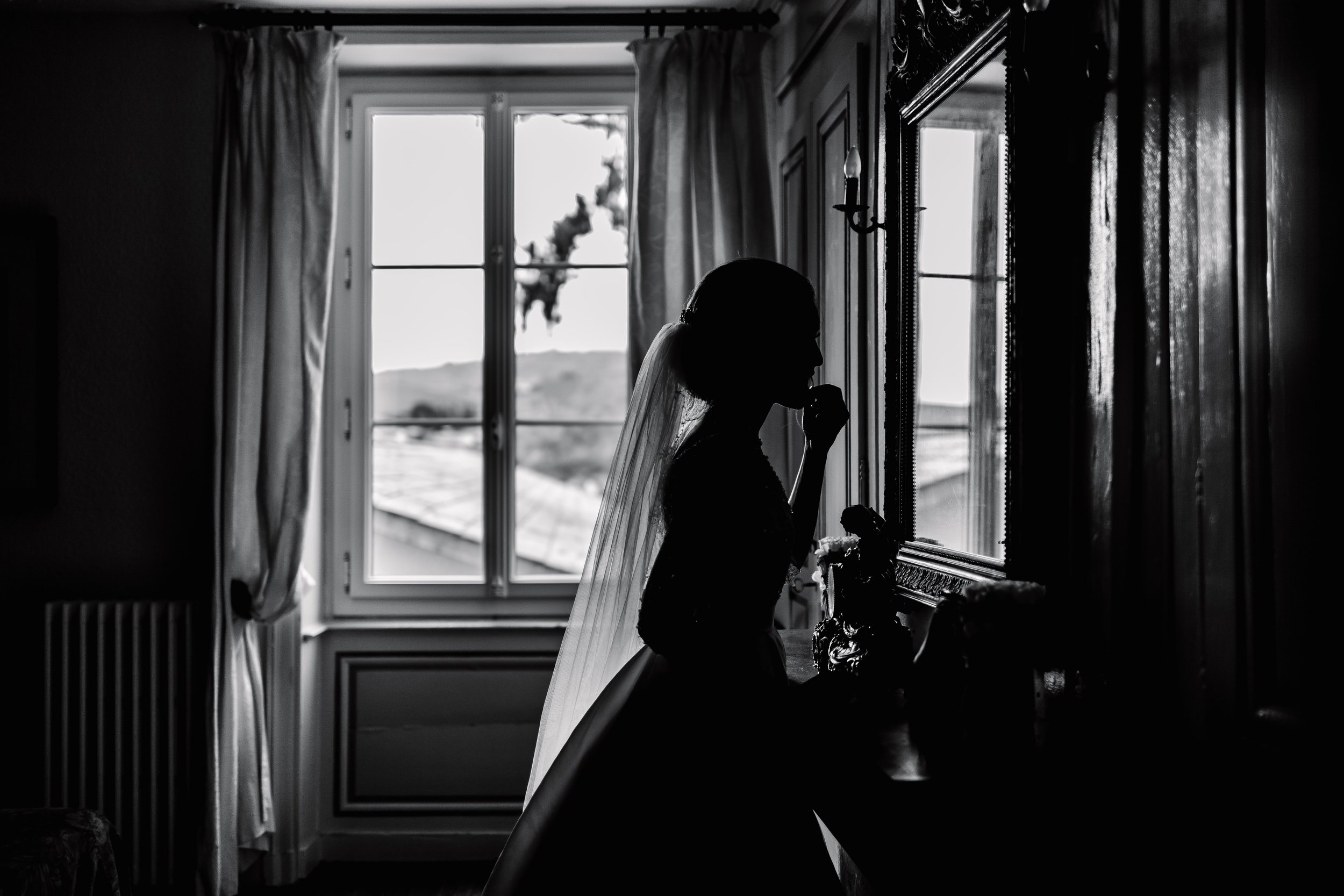 Silhouette of a bride wearing a veil, standing in front of a mirror inside a room with a window, curtains, and wooden wall panels.