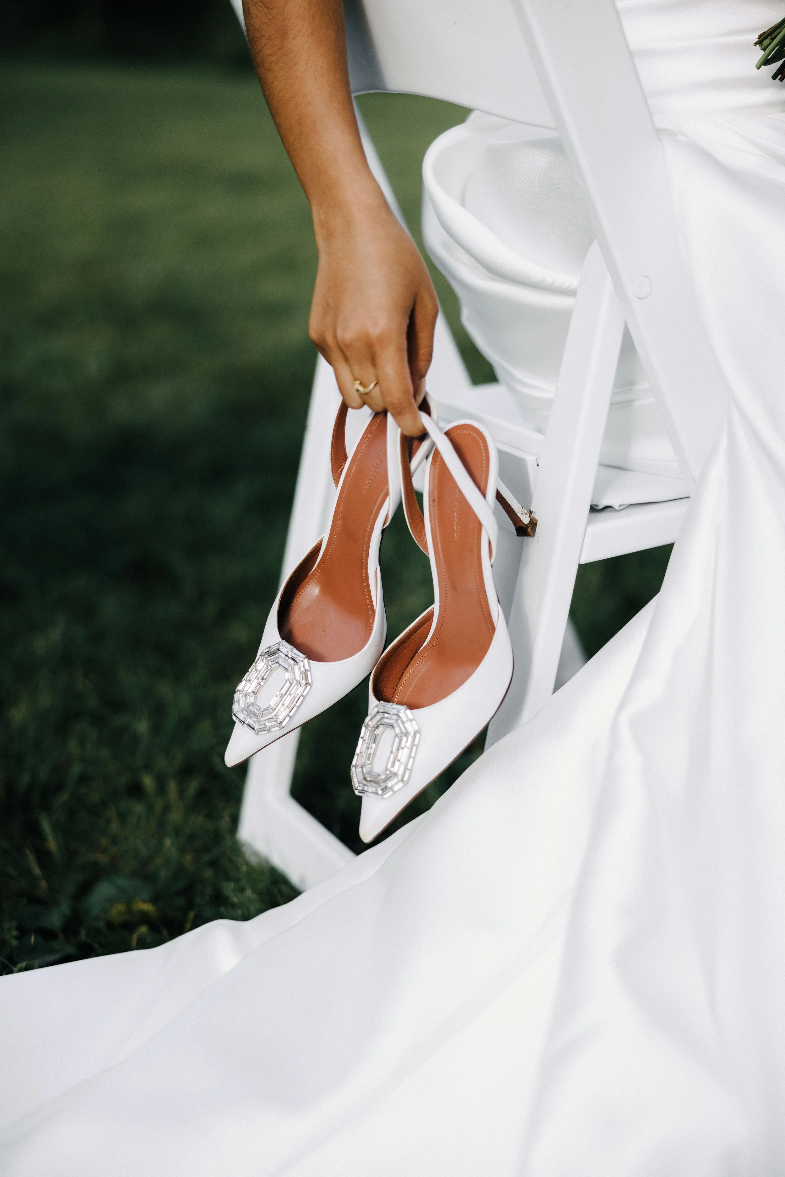 A person holding a pair of white high-heeled shoes with large rectangular rhinestone embellishments on the toes, sitting on a white chair on green grass, with a white dress partially visible.