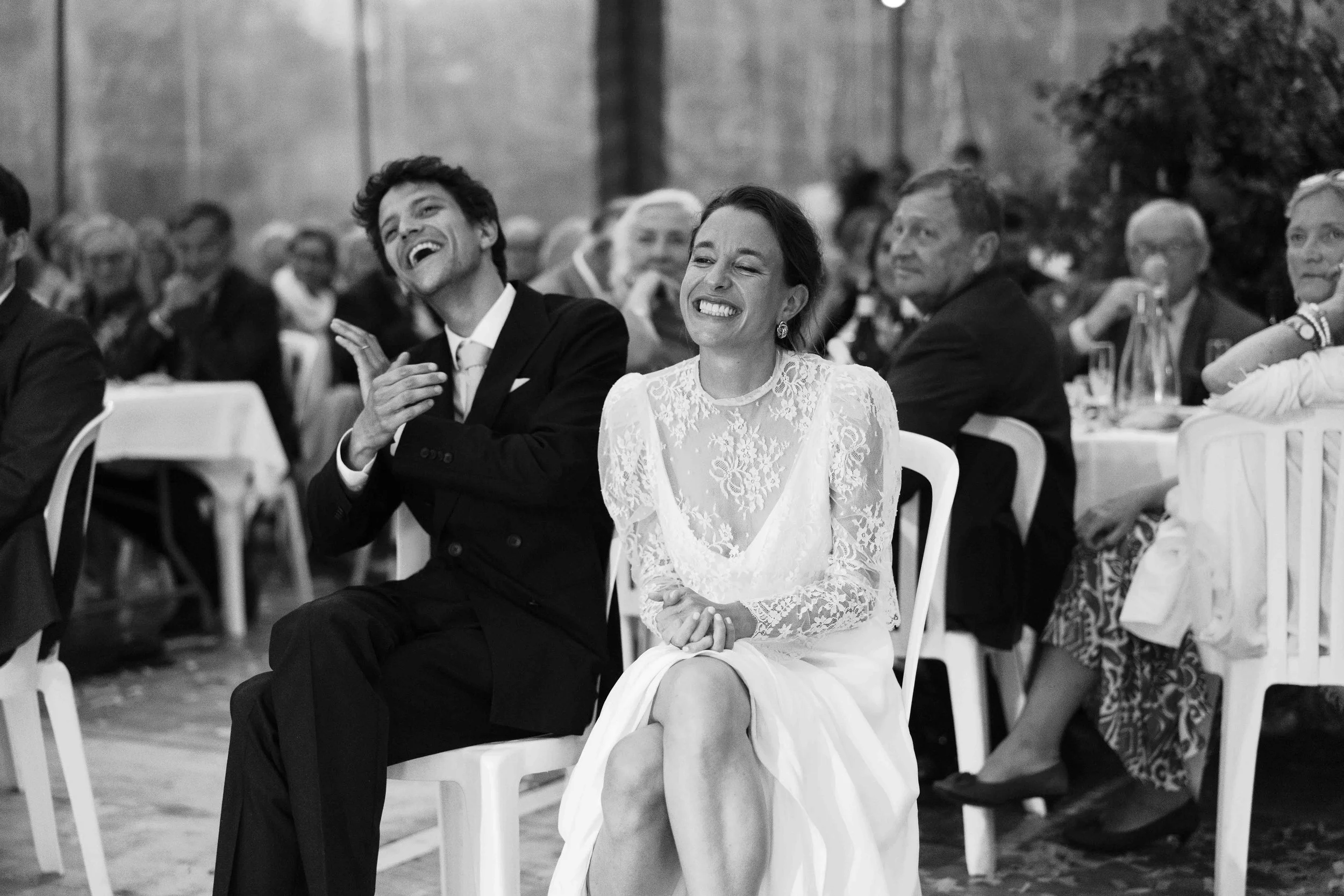 A bride and groom seated at a wedding reception, laughing happily while surrounded by guests.