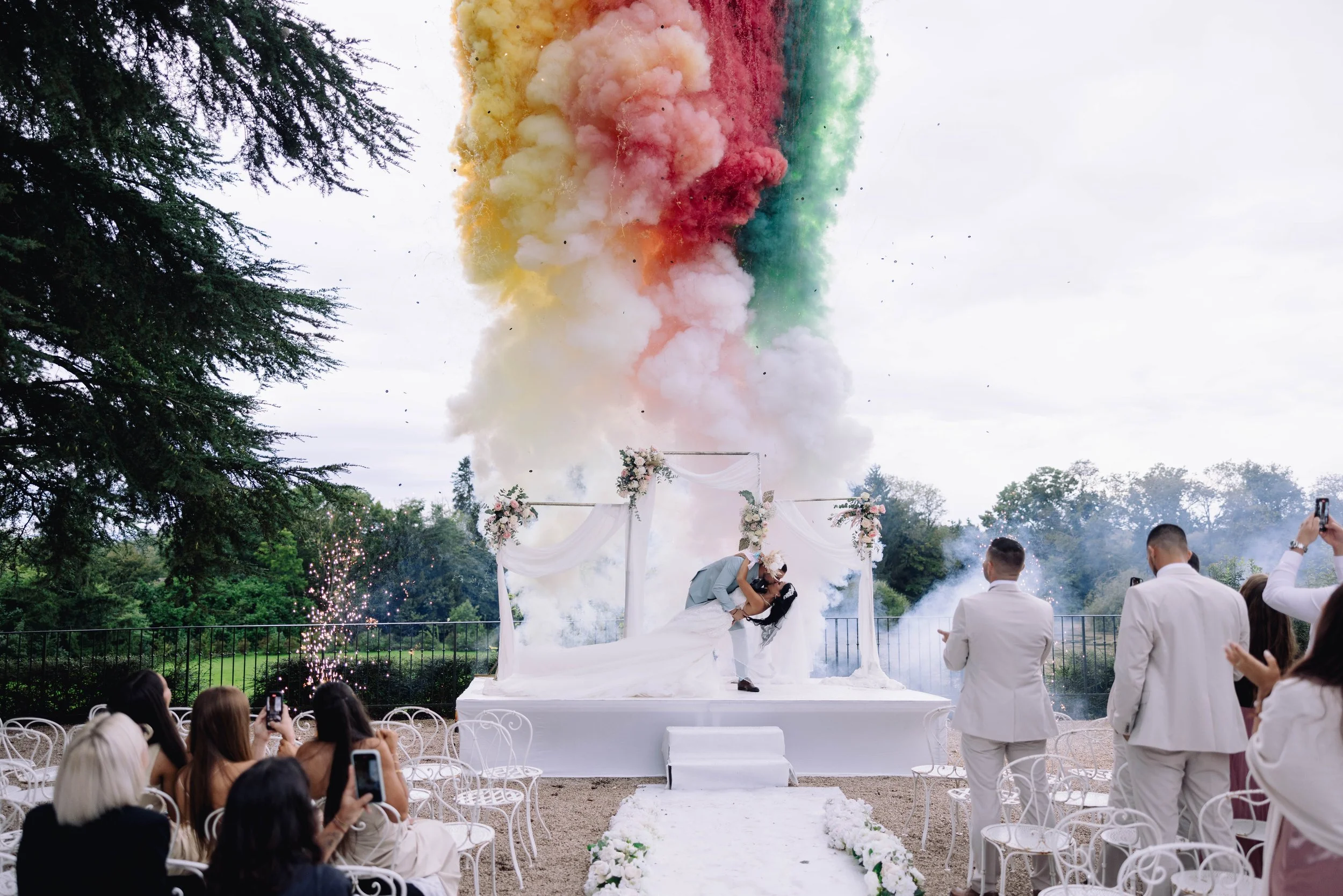 A wedding ceremony outdoors with a couple kissing on a decorated stage amid colorful smoke and fireworks, while guests capture the moment.