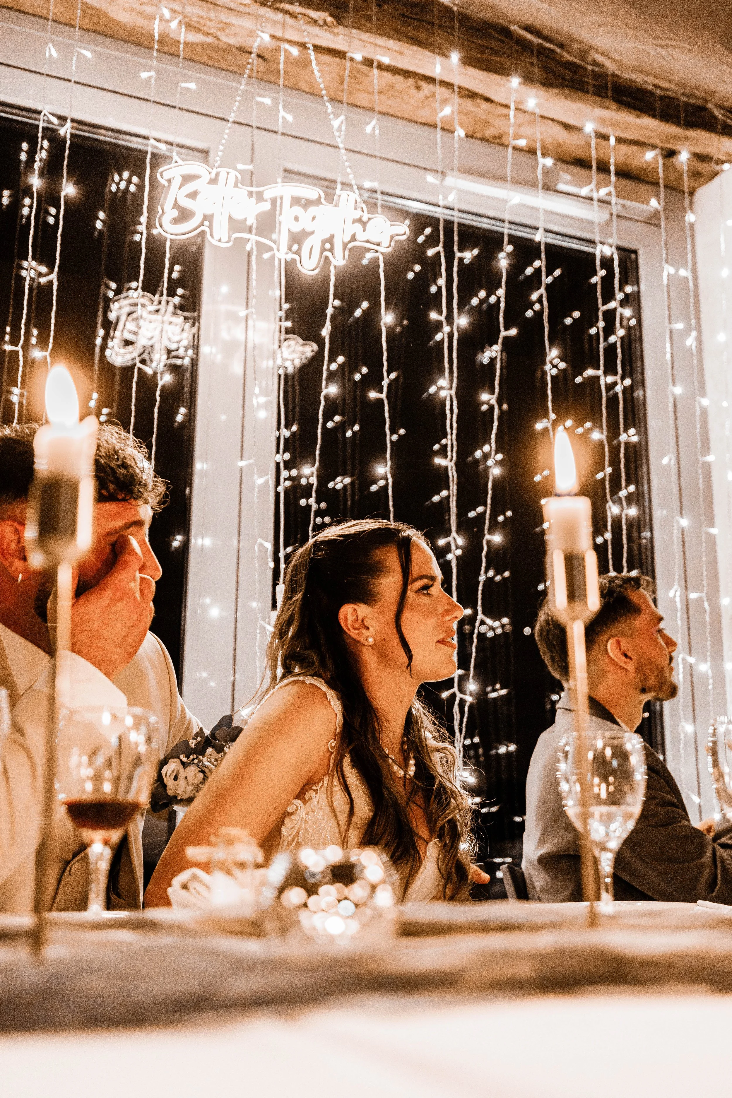 People sitting at a wedding reception table with lit candles and fairy lights, a woman in a wedding dress is smiling, celebrating indoors.